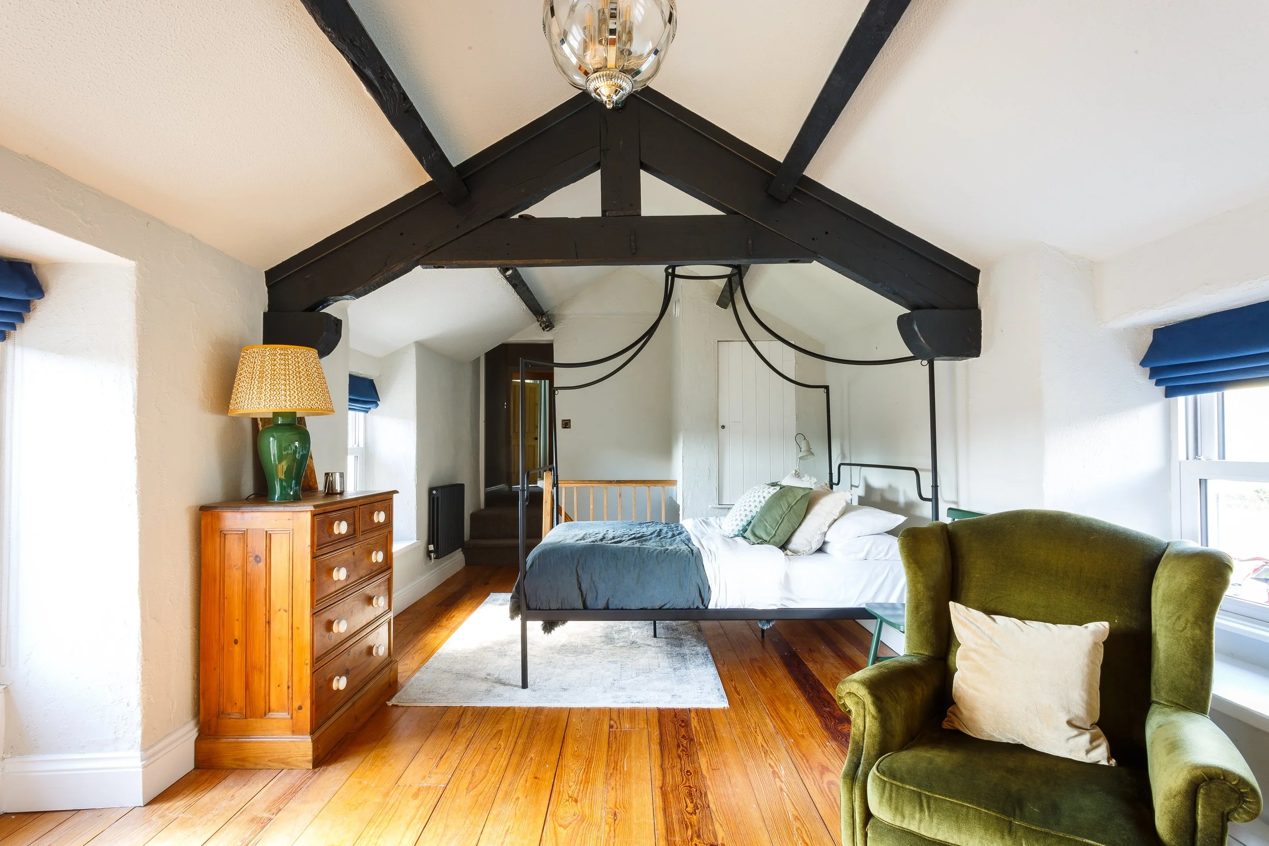 Light-filled attic bedroom featuring exposed black wooden beams, a wrought-iron bed frame, and polished wooden floors. The space is styled with vintage furniture, including a wooden chest of drawers, a green velvet armchair, and soft neutral textiles