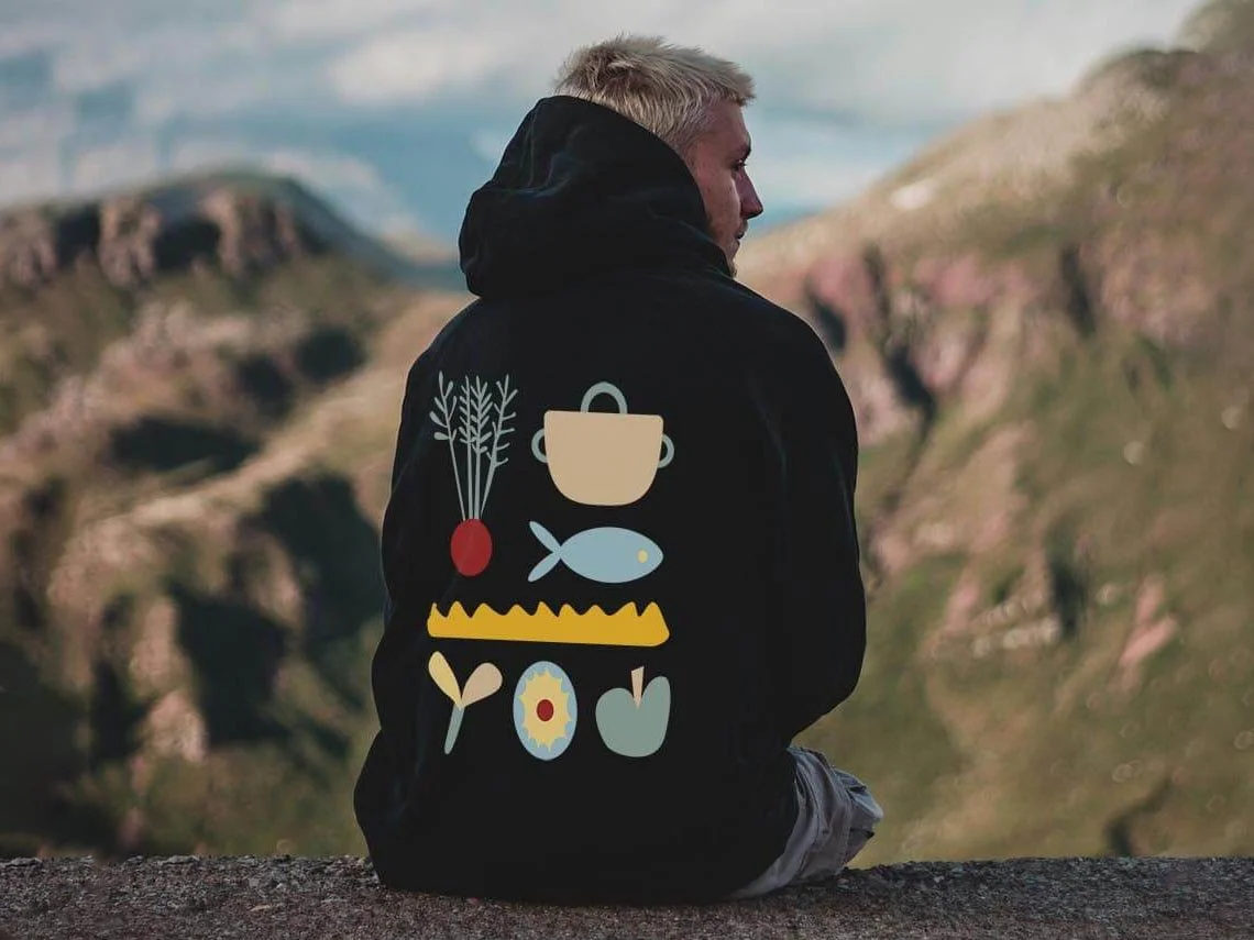 Eine Person mit blonden Haaren trägt einen schwarzen forikolo-Hoodie vor einer Bergkulisse. Die Illustrationen auf dem Hoodie sind eine Rübe, ein Topf, ein Fisch und ein Apfel.