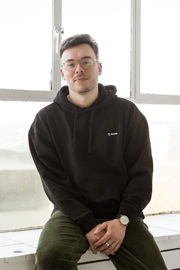 MarkOp Studio Ansprechpartner Alexander sitzt in einem schwarzen Pullover mit MarkOp Logo auf einer weißen Fensterbank.
