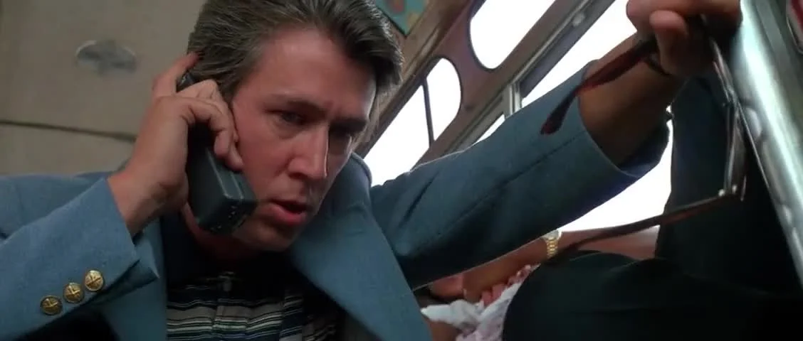 Man with brown hair speaking on a landline phone inside a vehicle, reaching with his hand towards the dashboard.