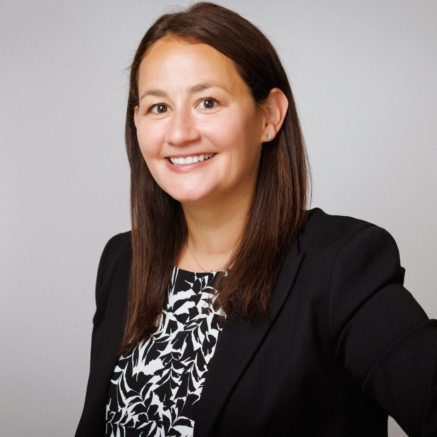 A portrait of a woman with long brown hair wearing a black blazer with a patterned black and white top, smiling against a plain gray background.