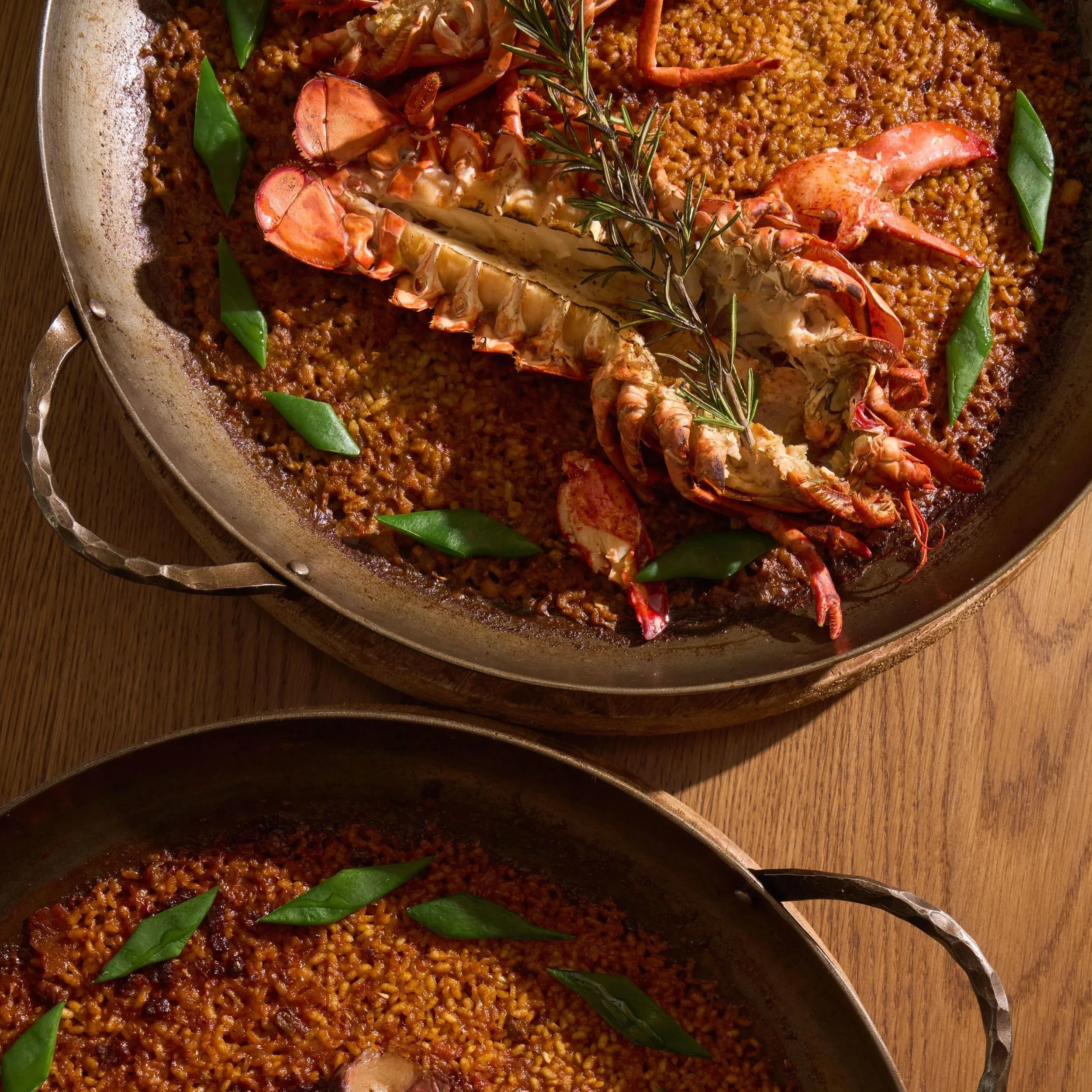 Close-up of a seafood paella with a lobster on top, garnished with fresh rosemary and green vegetables in a large pan.