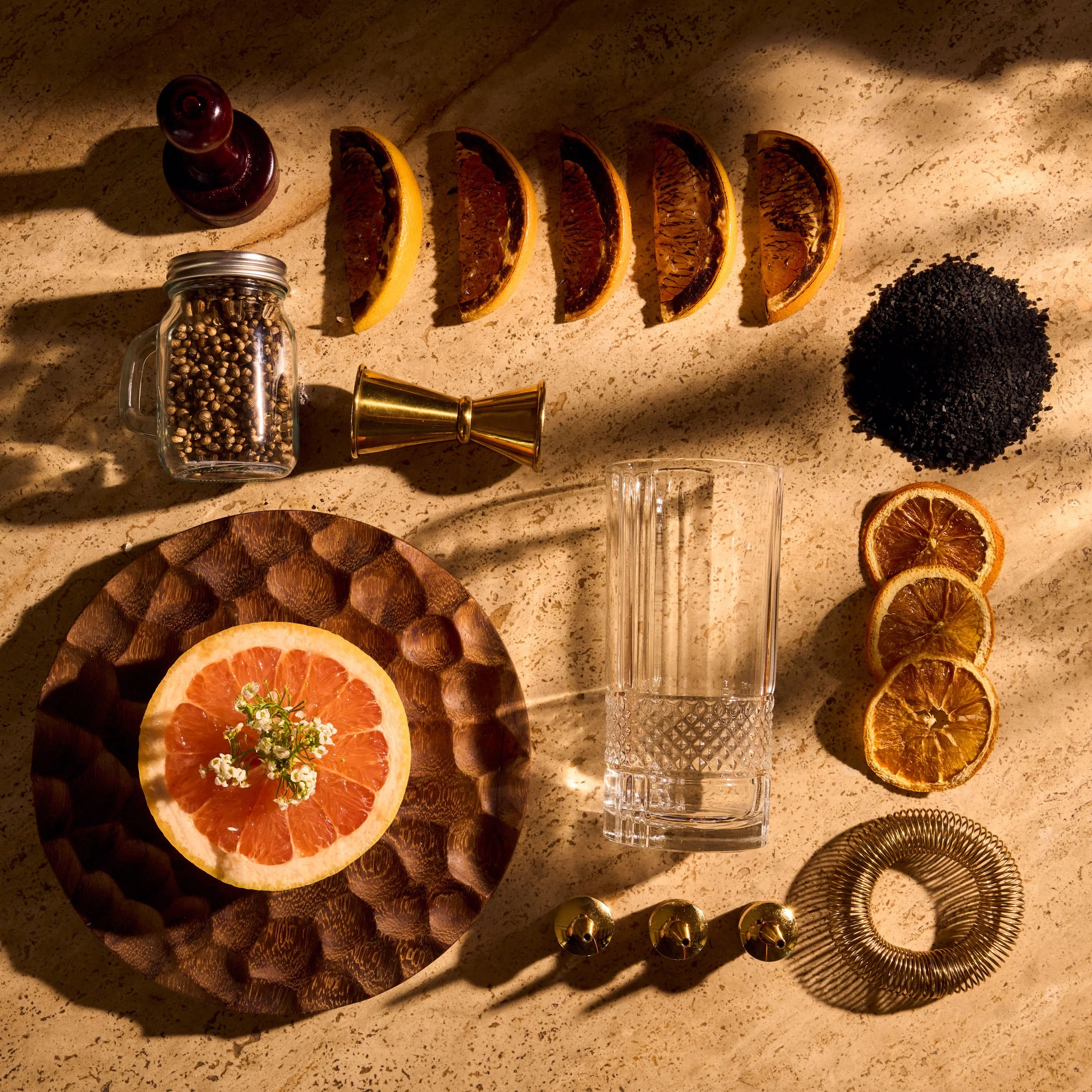 Slices of dried orange arranged on a dark wooden tray, with a small white flower and green herbs garnish. Nearby, there are black teabags, a glass jar of coriander seeds, a clear drink glass, a gold cocktail shaker, and various golden bar tools on a beige speckled countertop.