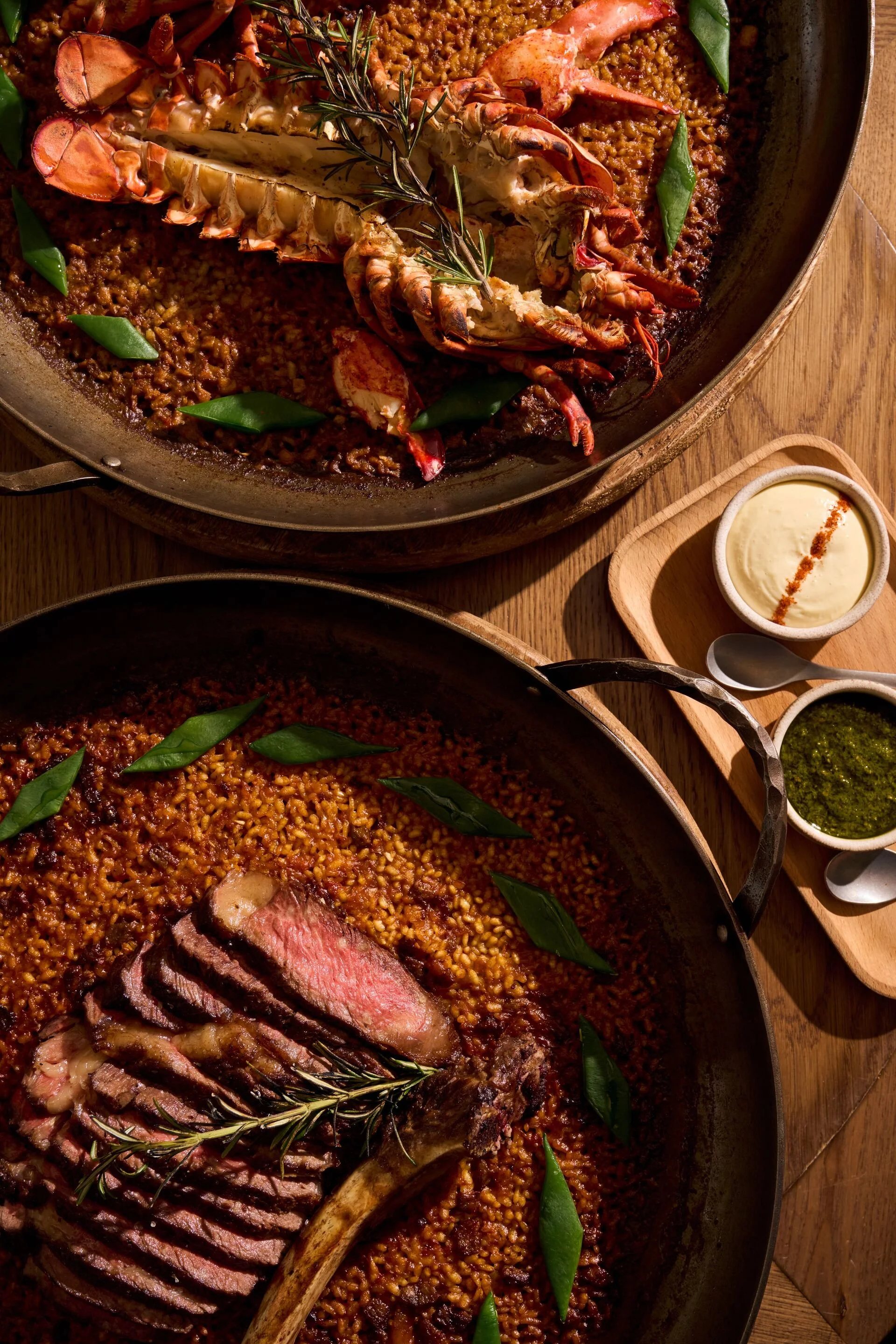 A large dish of cooked rice or grains with slices of roasted steak, garnished with green leaves, alongside a lobster or crayfish, and some sprigs of rosemary, served with dipping sauces.