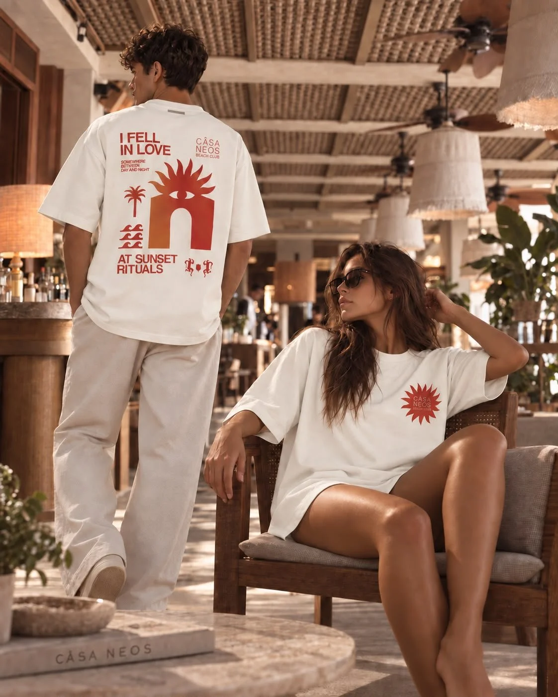 A man and a woman in casual clothing inside a rustic, tropical-themed cafe. The man stands with his back to the camera, wearing a white T-shirt with red graphic text and designs and beige pants. The woman sits on a wooden chair with her legs crossed, wearing a white T-shirt with red graphic, sunglasses, and has wavy brown hair.