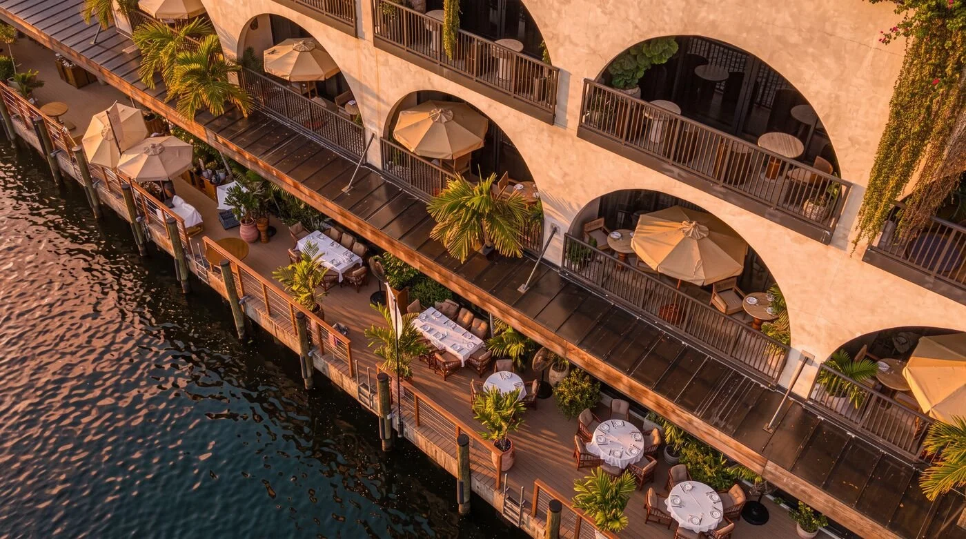 An aerial view of a lakeside restaurant with outdoor dining tables, potted plants, and umbrellas, adjacent to a building with arched balconies.