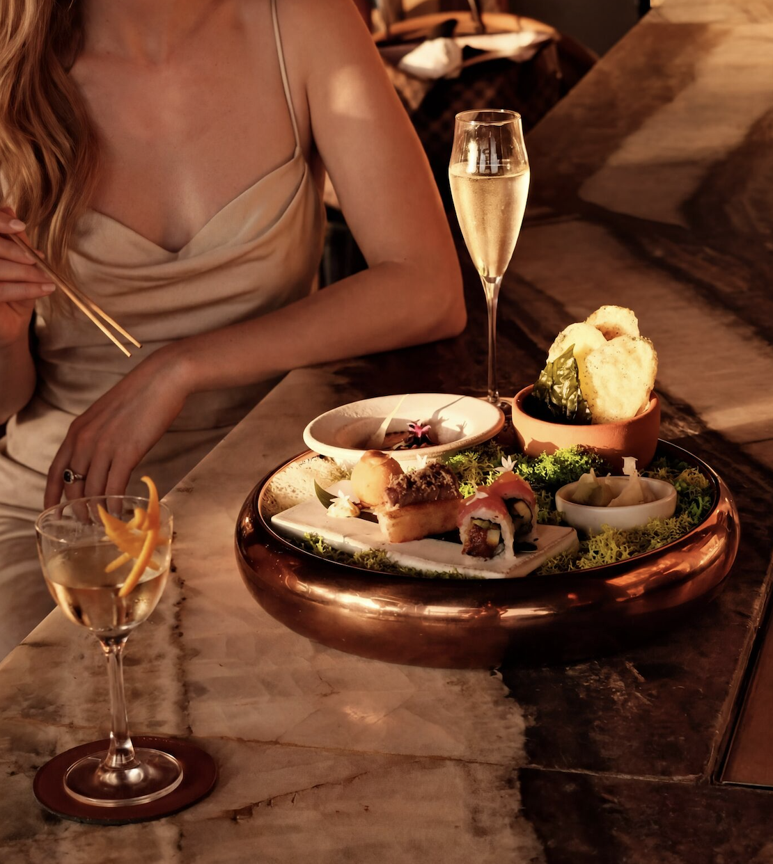 An elegant woman is at the bar of CASA NEOS for golden hour, having a cocktail and a selection of bites from the menu, including a dip, sushi, and more.