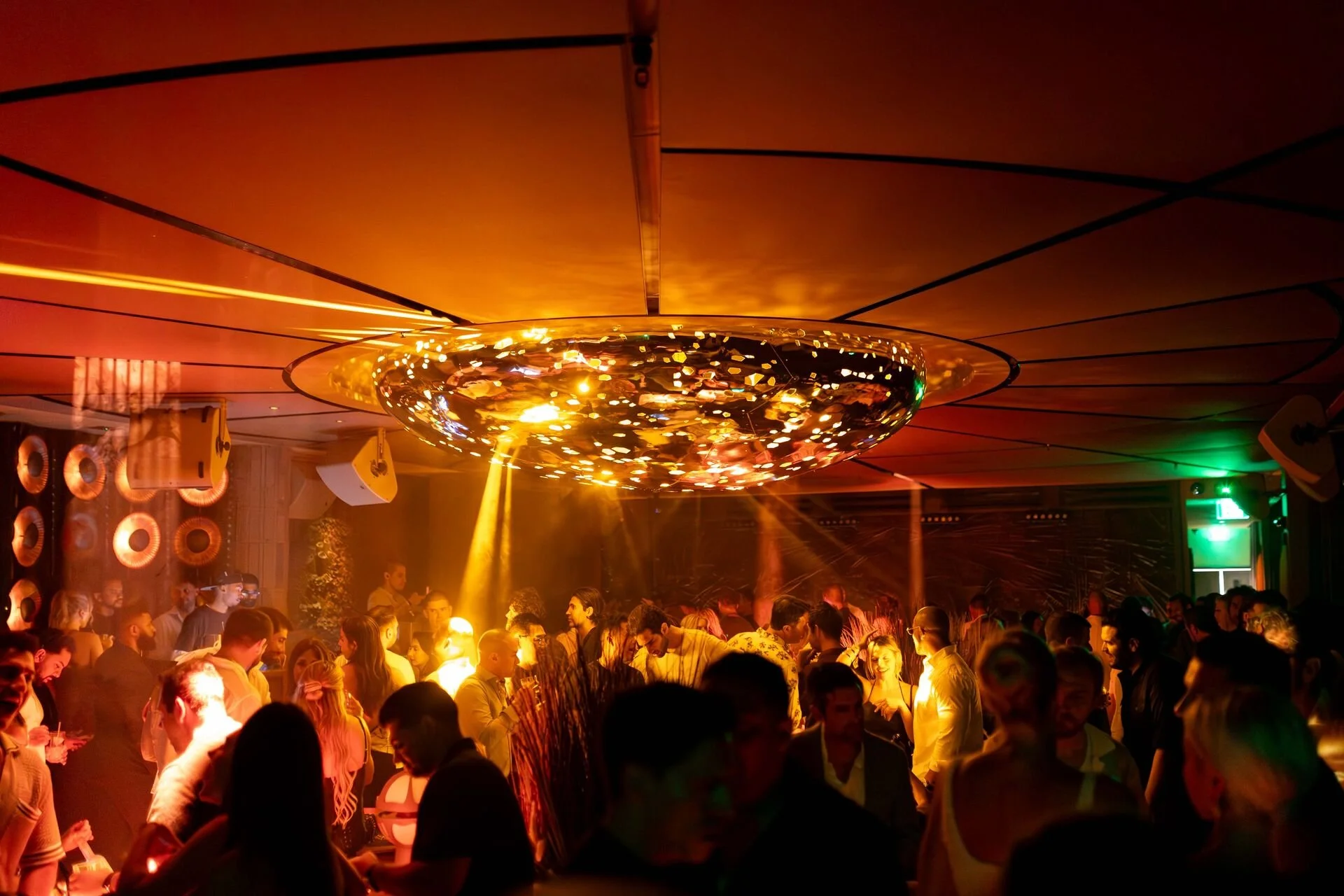Crowd of people dancing and socializing in a nightclub with colorful lighting and a large pendant light fixture hanging from the ceiling