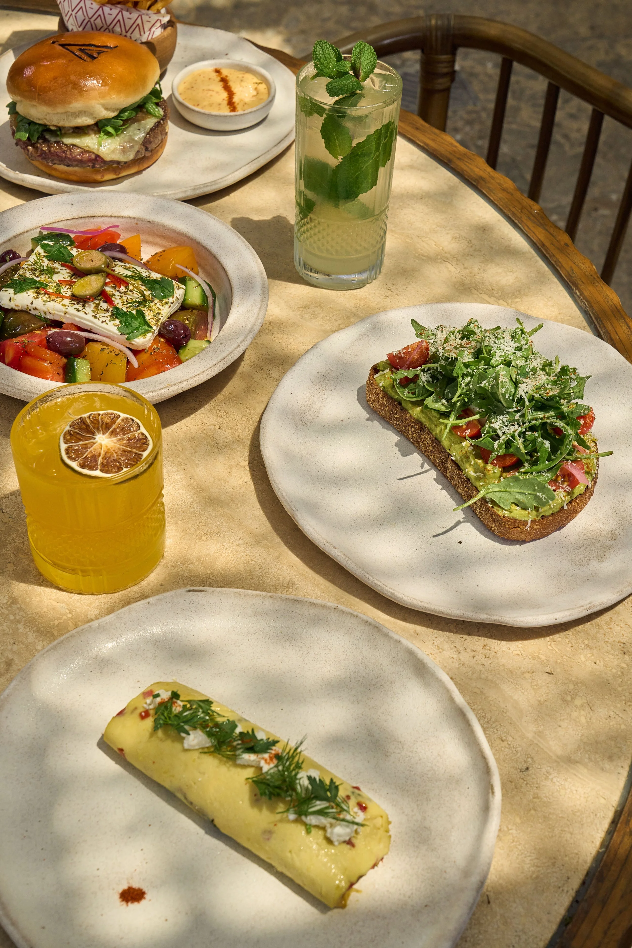 A brunch spread of CASA NEOS dishes, including the Greek omellete, avocado toast, greek salad, and a burger with two refreshing cocktail drinks.
