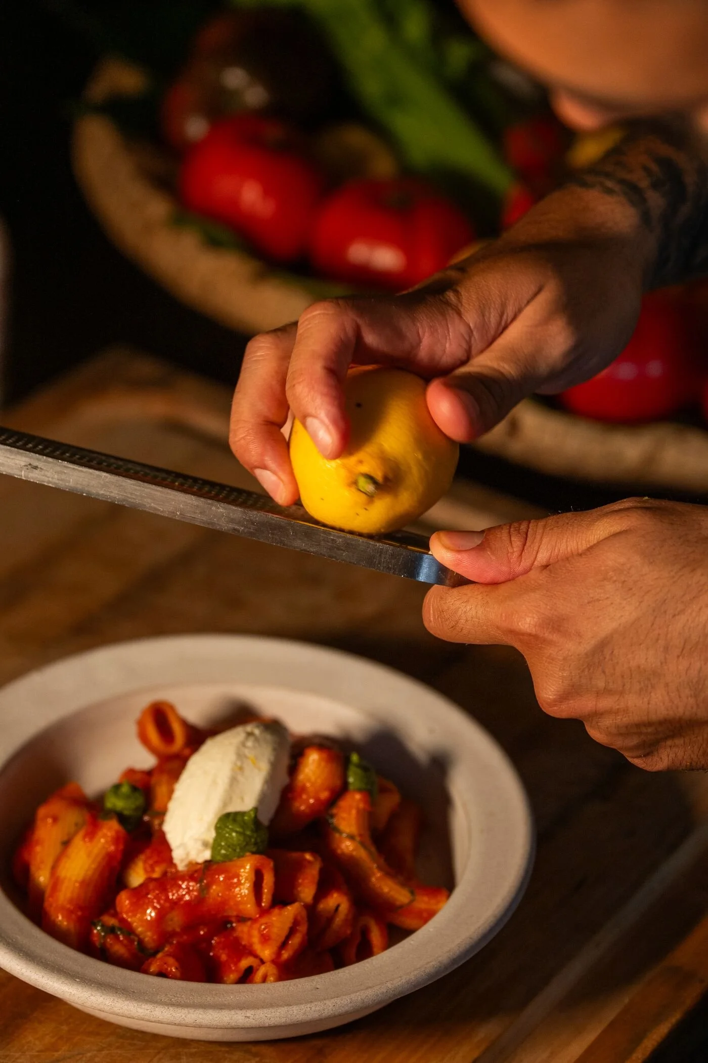 A person is holding a lemon above a bowl of food, with a variety of chopped vegetables in the background. The bowl contains pasta with red sauce, garnished with a slice of mozzarella cheese and green herbs.