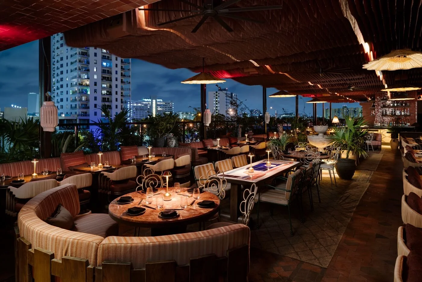 Nighttime view of an upscale rooftop restaurant with round and rectangular tables, elegant chairs, and a city skyline with tall buildings in the background.