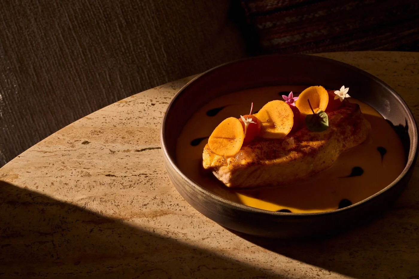 A bowl of dessert with a pastry garnished with banana slices and edible flowers, served on a textured surface.