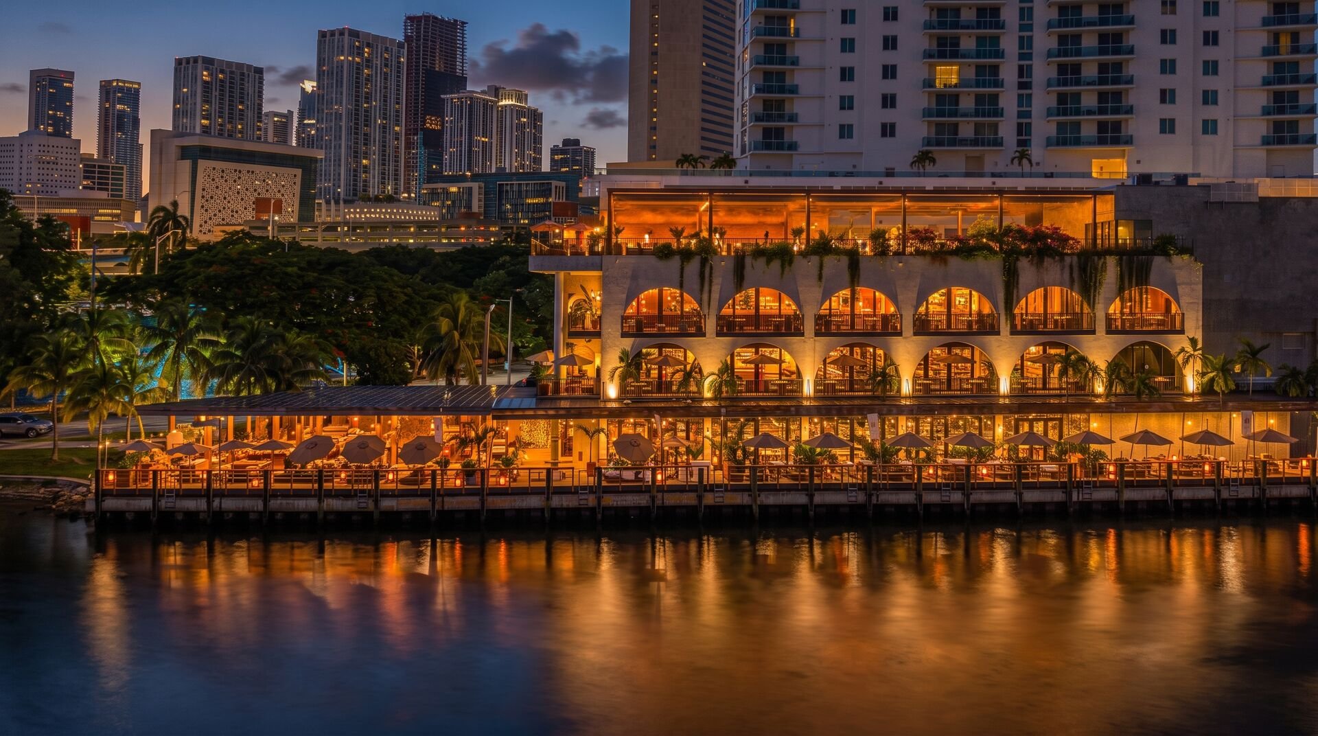 Waterfront restaurant glowing at dusk with palm trees and a high-rise city skyline in the background
