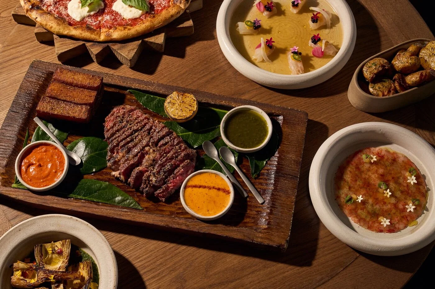 An assortment of Indian dishes, including a grilled meat platter with sauces, a pizza, and various bowls of curries and dips on a wooden table.