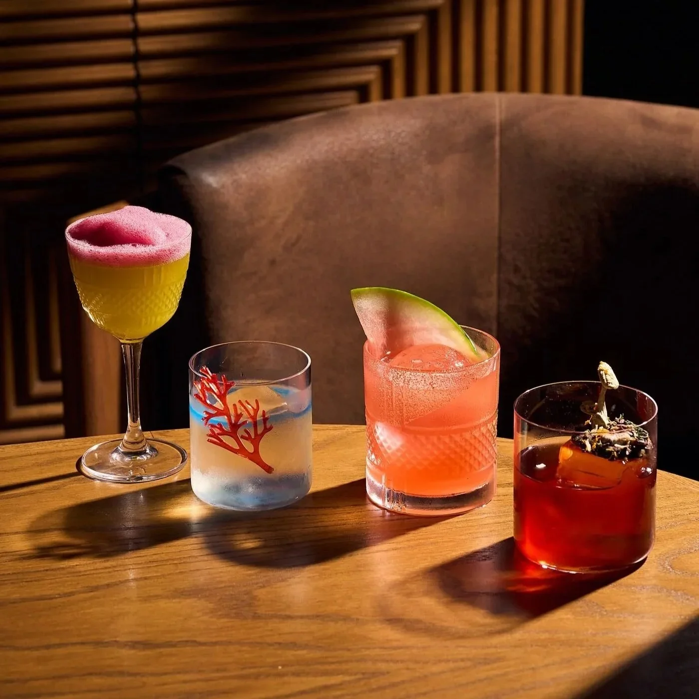 Four colorful cocktails on a wooden table, each in a different glass, with a brown leather chair and wooden wall in the background.