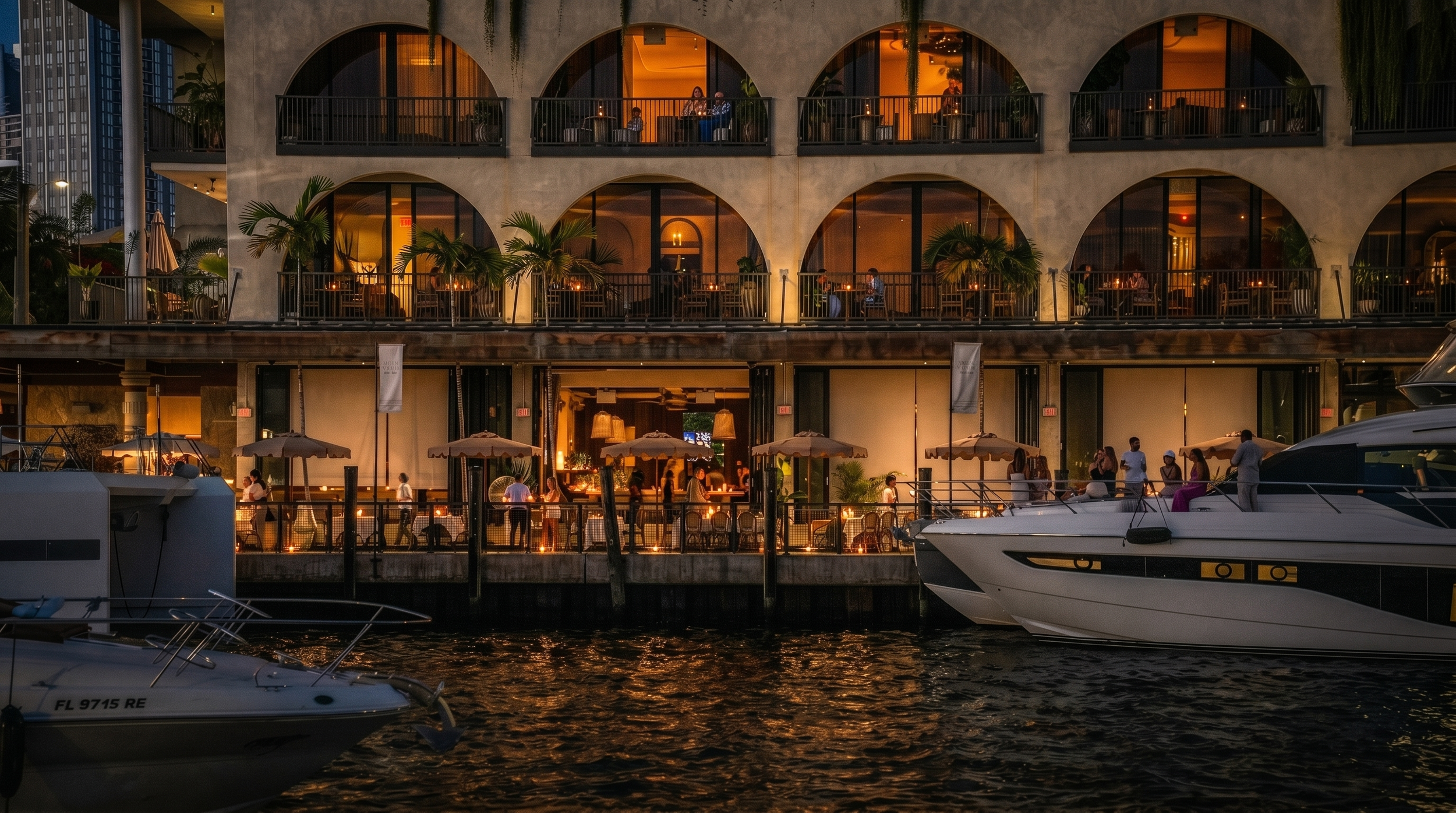 Night view of a multi-story waterfront restaurant with outdoor dining on each level, illuminated by warm lighting, with palm trees and people dining and socializing, alongside boats docked in the water.