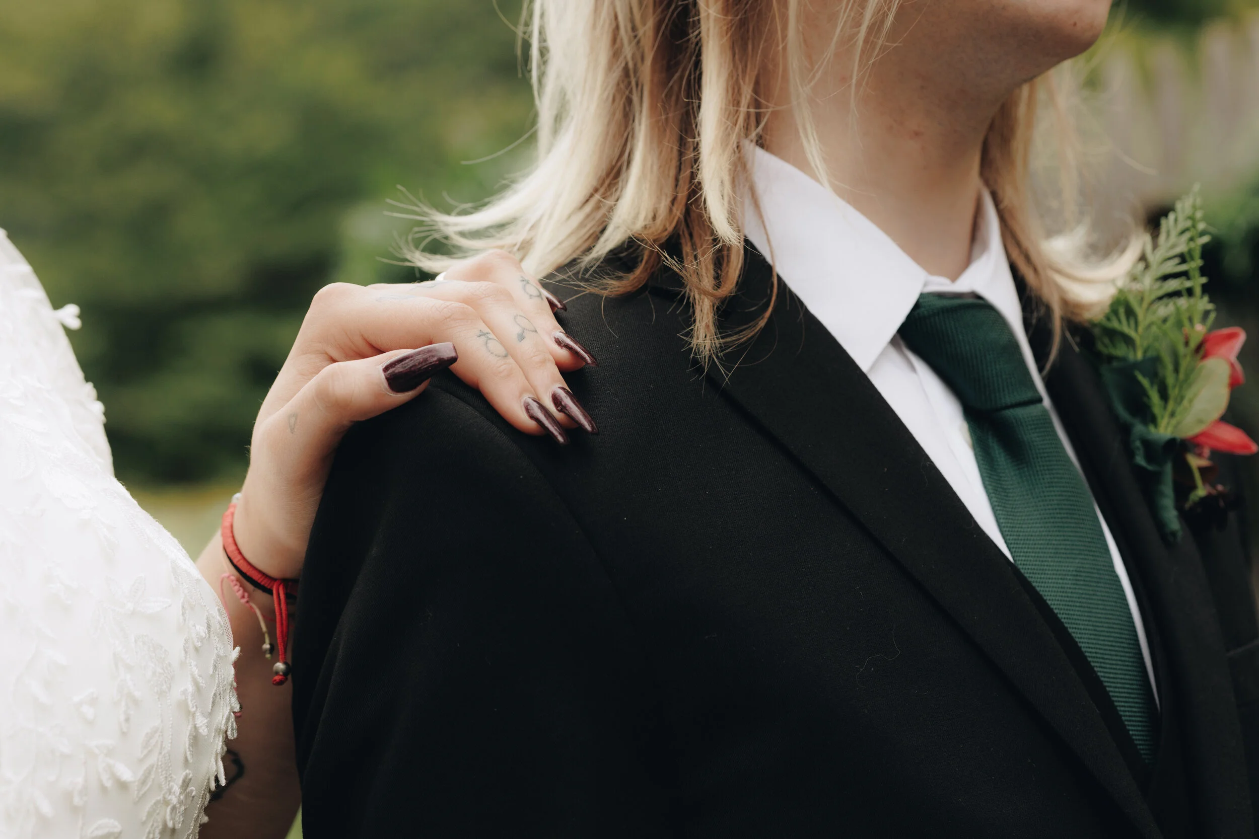 Close-up of a woman in a black tuxedo with a white shirt and green tie, with a boutonnière pinned to her lapel, and a hand with dark nail polish resting on her shoulder.