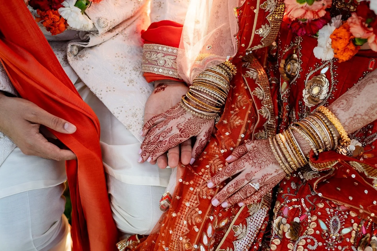 Close-up of a traditional Indian wedding ceremony showing a bride and groom holding hands. The bride is dressed in a red and gold embroidered sari and adorned with gold jewelry and mehndi on her hands. The groom is wearing white and orange garments with a red dupatta, and a hand with henna touches the bride's hand.