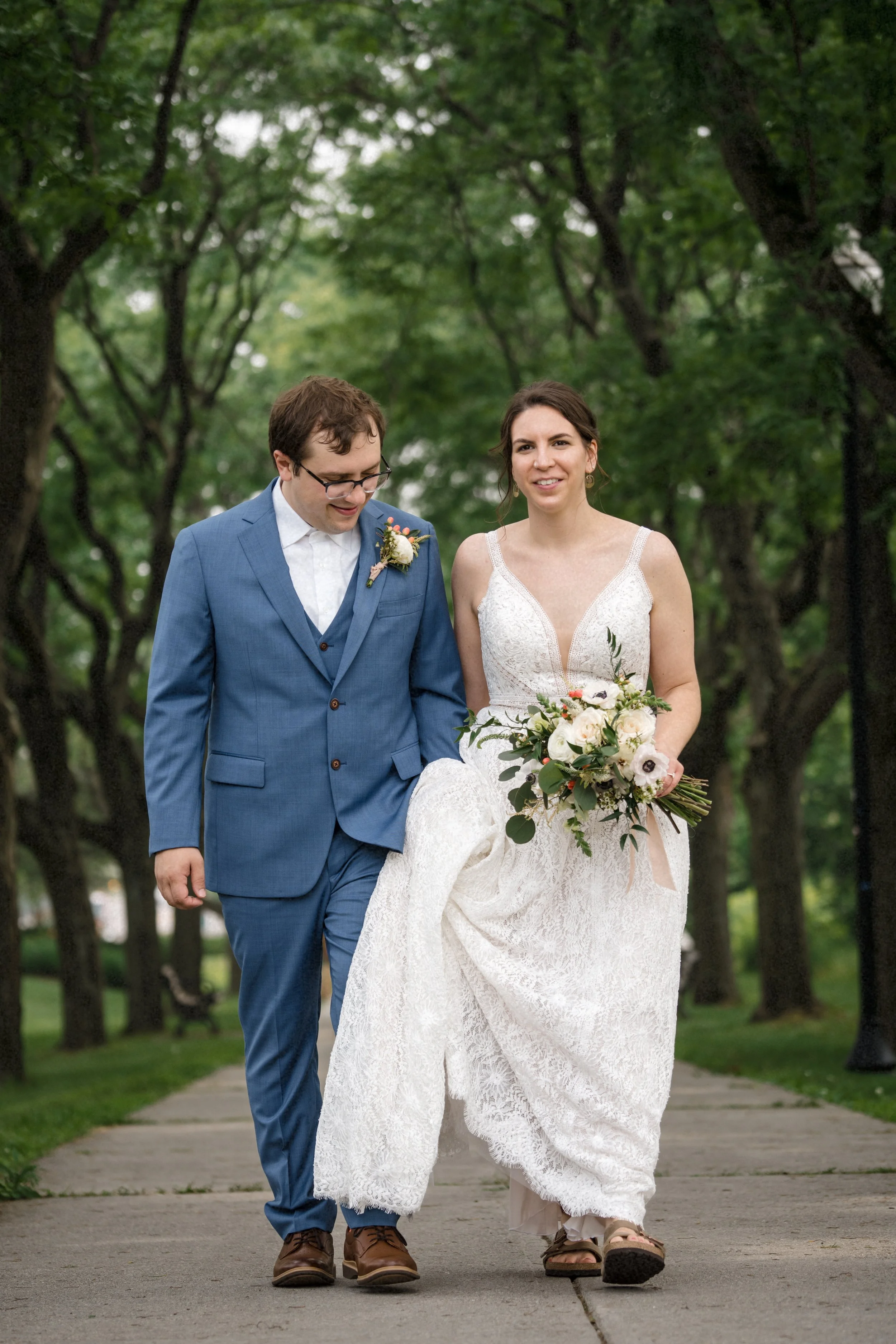 A bride and groom walking together in a park, the bride in a white lace wedding dress holding a bouquet, and the groom in a blue suit with a white shirt, during a wedding ceremony.