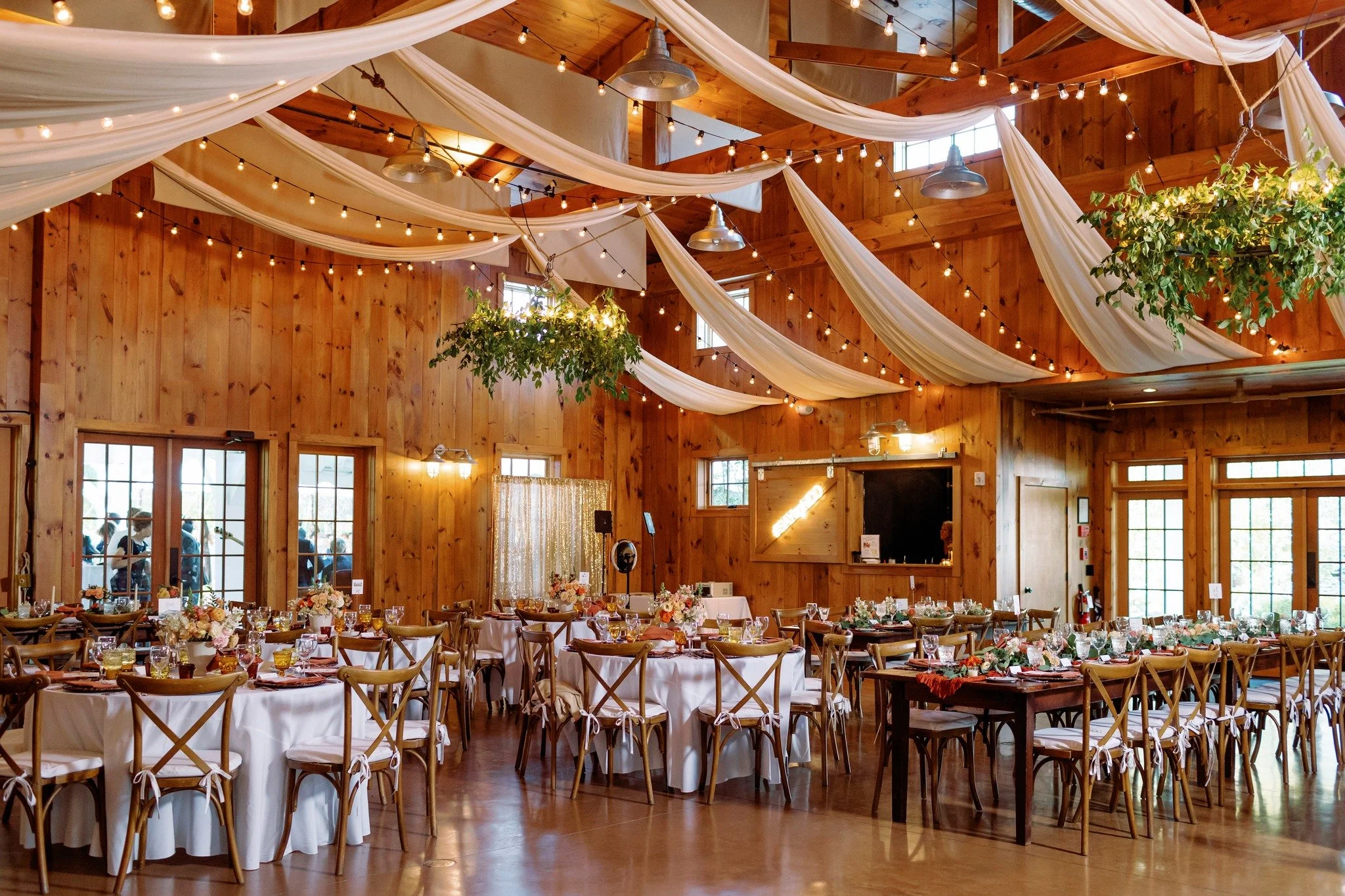 A decorated wooden event hall with draped white fabric and string lights hanging from the ceiling, set up with multiple round and rectangular tables with floral centerpieces, glassware, and tableware for a celebration or wedding reception.