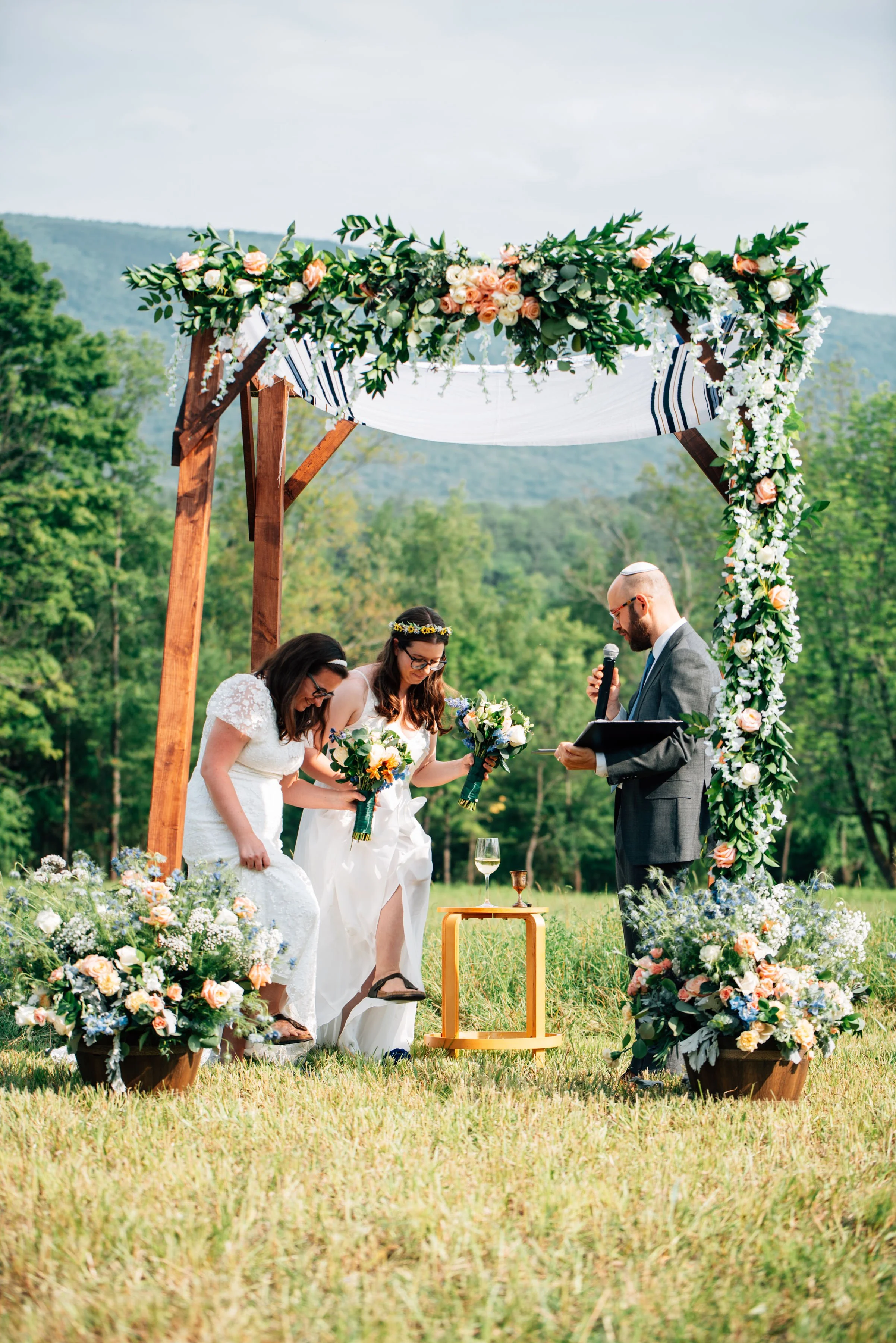 Outdoor wedding ceremony with two brides and an officiant under a floral arch, surrounded by greenery and flowers.