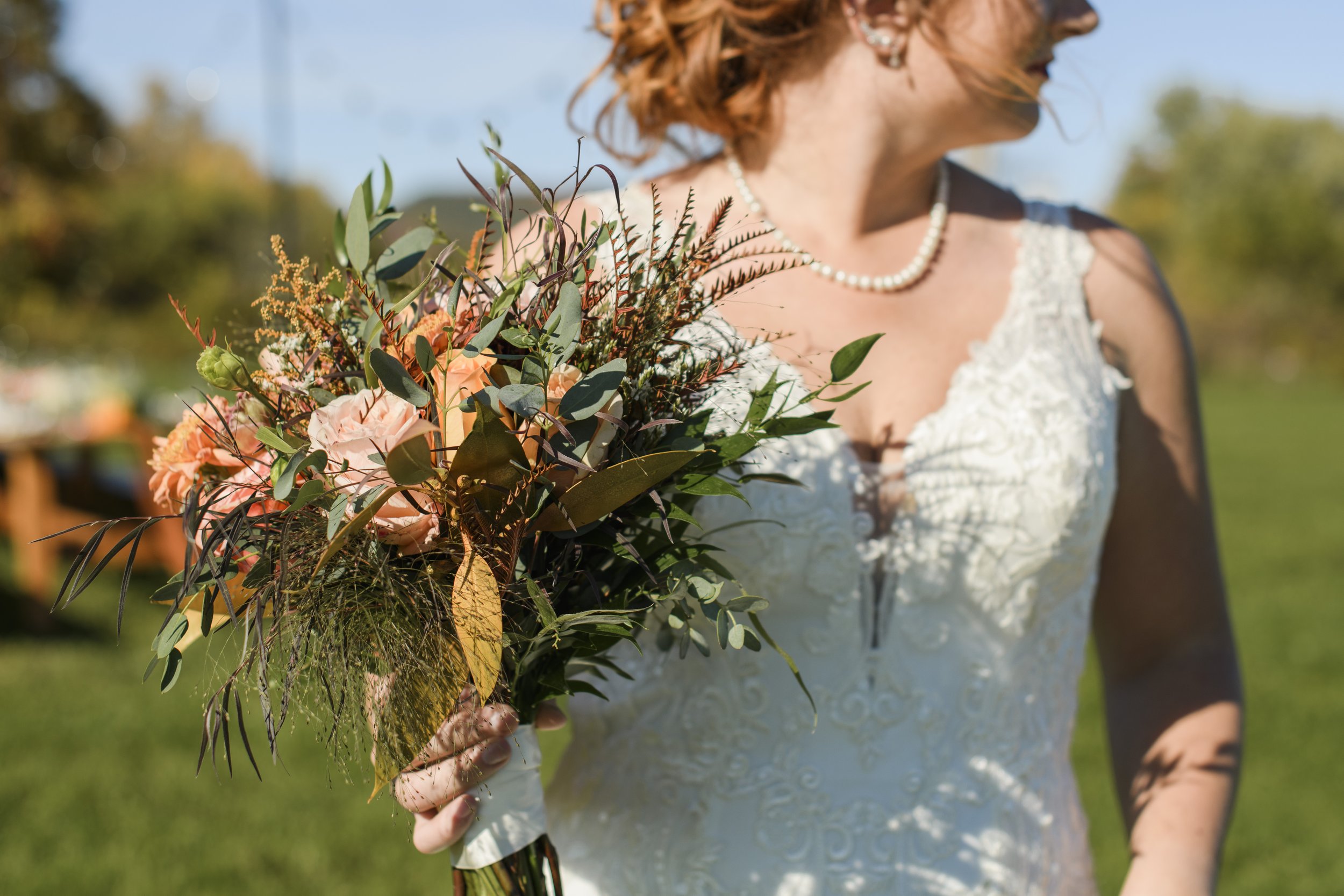 Woman in wedding dress holding a bouquet of pink and orange flowers with greenery, outdoors on a sunny day.