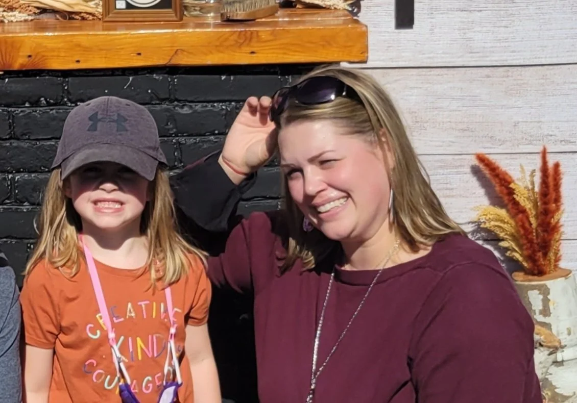 A woman and a young girl smiling outside, with the woman touching sunglasses on her head, in front of a black brick wall and white wooden siding.