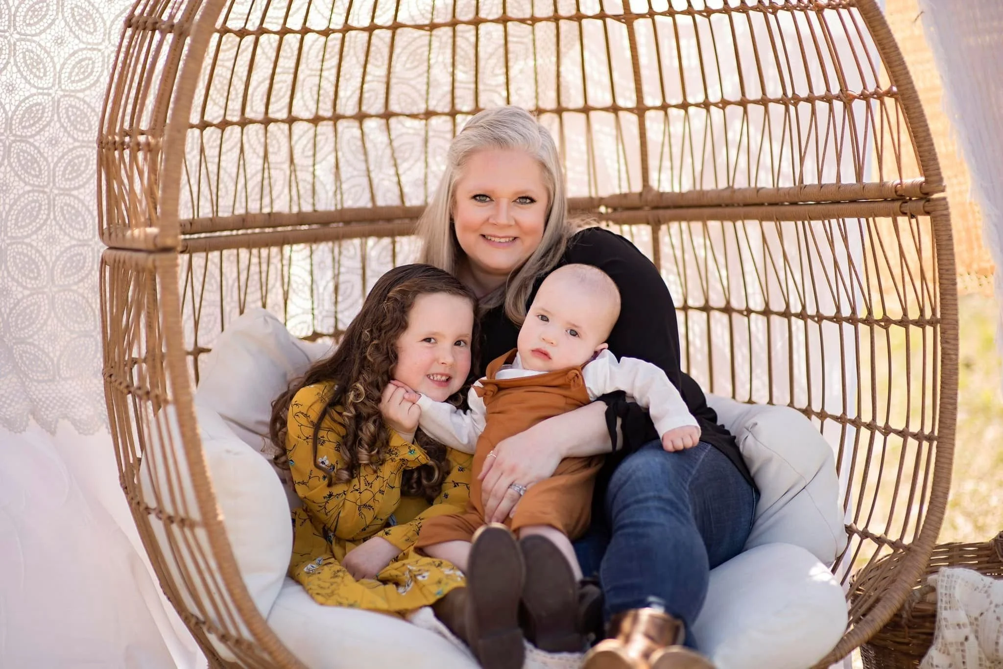 A woman sitting inside a hanging wicker egg-shaped chair with two children, a girl with curly hair wearing a yellow dress and a boy with a bald head in brown overalls. They are smiling and posing for the photo.