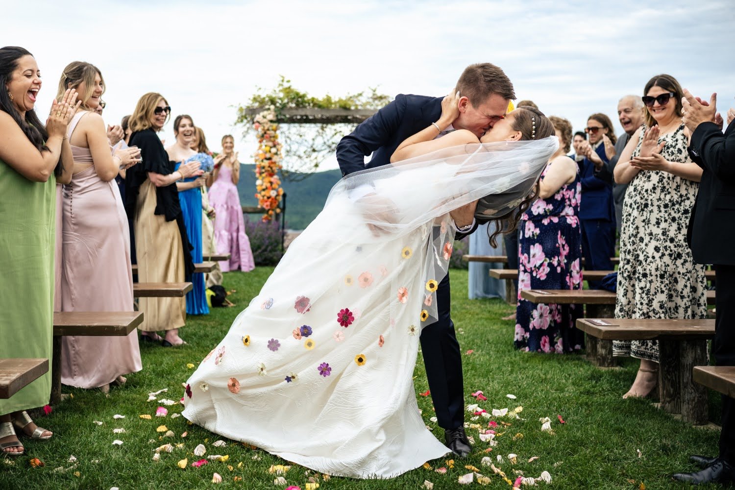 Bride and groom kissing while bending at a wedding ceremony, surrounded by clapping guests on a grassy outdoor setting.