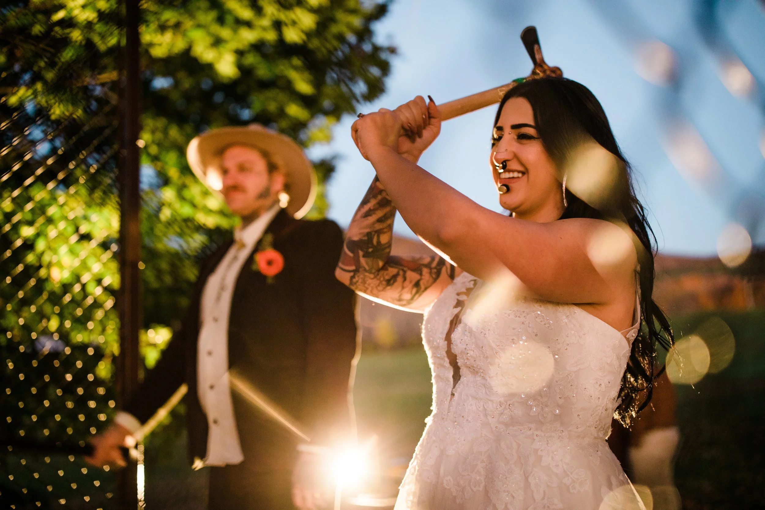 A woman with dark hair, tattoos, and piercings smiling and holding an axe over her shoulder at an outdoor evening event, with a man in a suit and hat in the background.