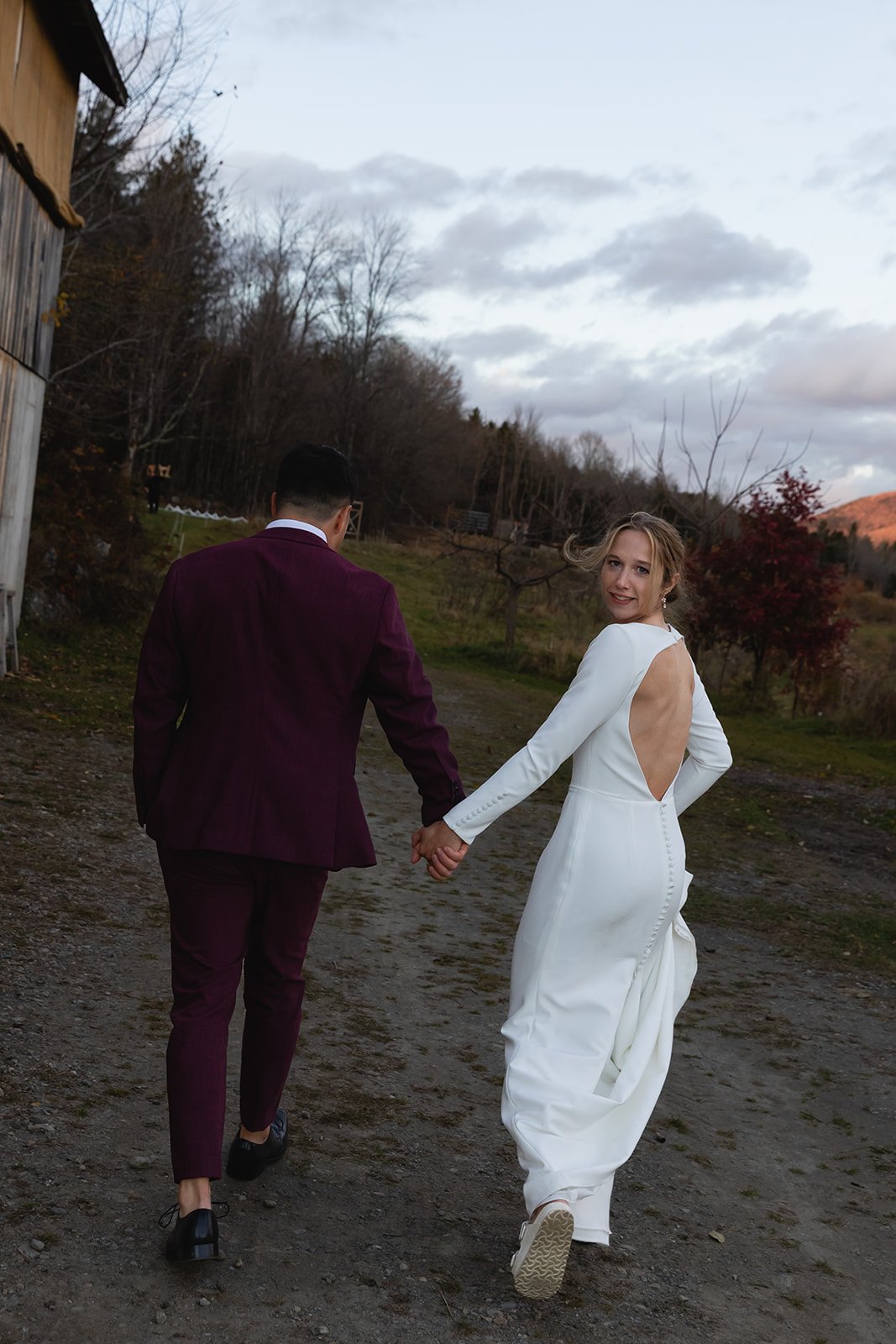 A bride in a white wedding gown and a groom in a dark suit holding hands outdoors on a dirt path, with trees and a cloudy sky in the background.