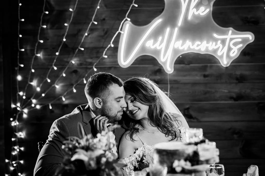 A couple sitting at a table with a wedding cake, smiling and leaning their foreheads together at their wedding reception.
