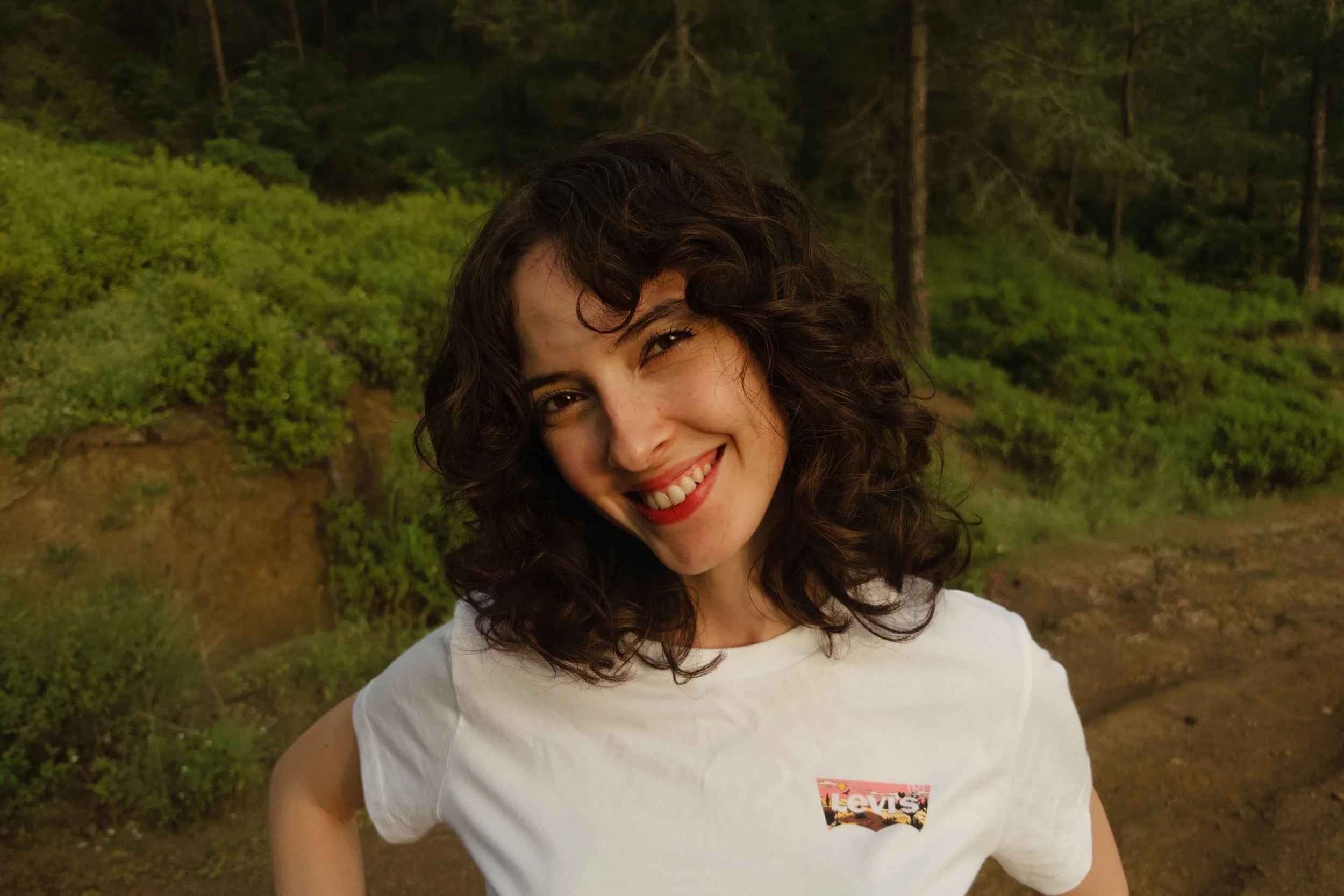 A woman with curly dark hair smiling outdoors in a forested area, wearing a white T-shirt with a Levi's logo. Noema Therapy in Toronto, Individual and Couples Therapy in Ontario