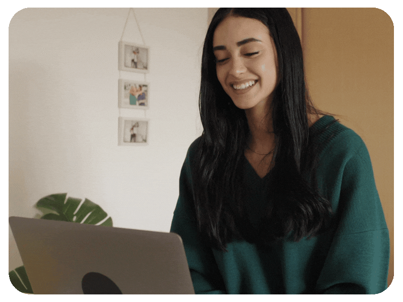A woman with long black hair smiling while looking at a laptop in a cozy indoor setting.