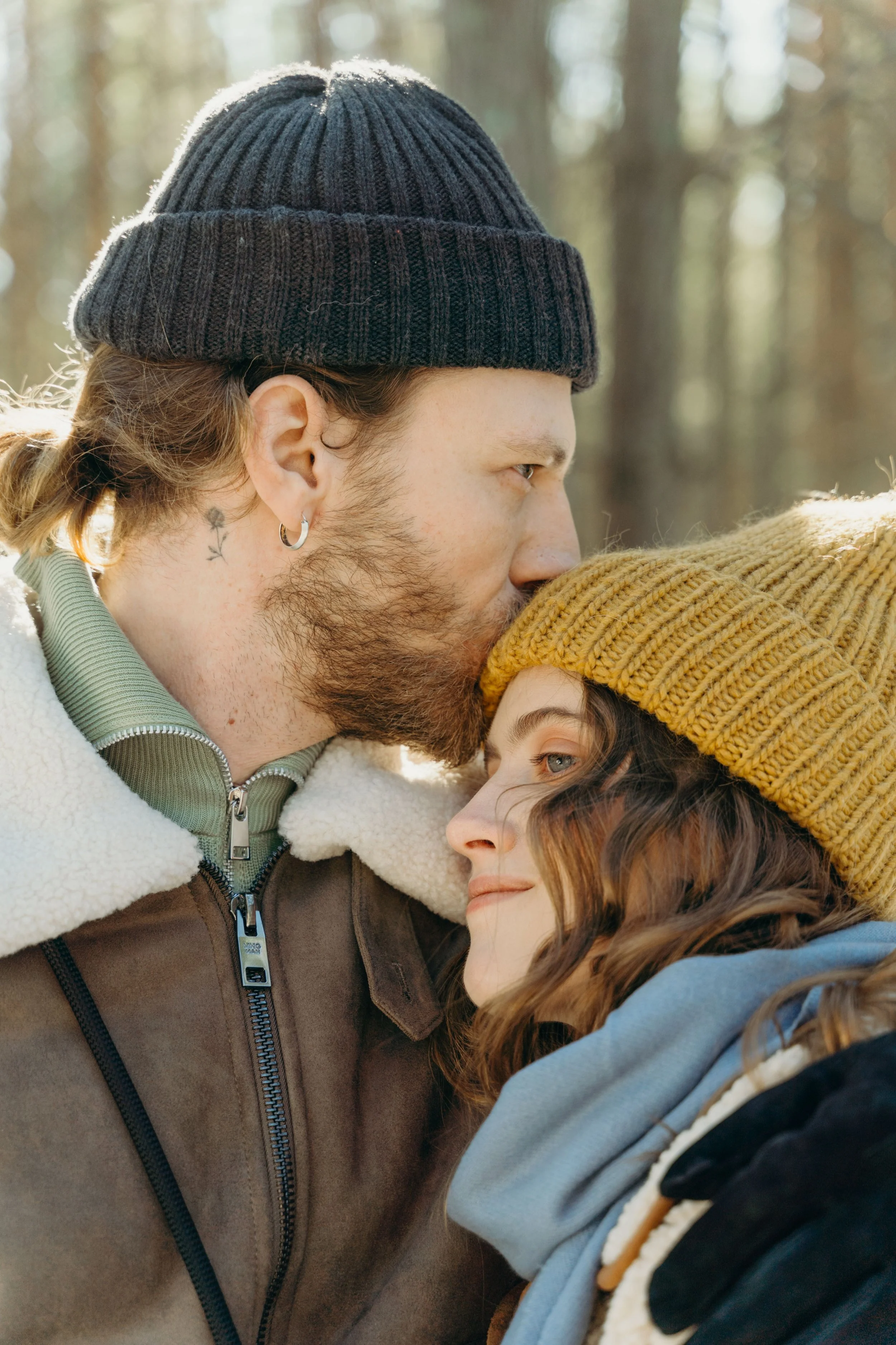 A man and a woman are close together, sharing a tender moment outdoors in a wooded area with trees in the background. The man is wearing a black knit beanie and a brown and cream fleece jacket. He has a small tattoo near his ear and an earring in his left ear. The woman is wearing a yellow knit beanie and a blue hoodie, and they are looking into each other's eyes with smiles.