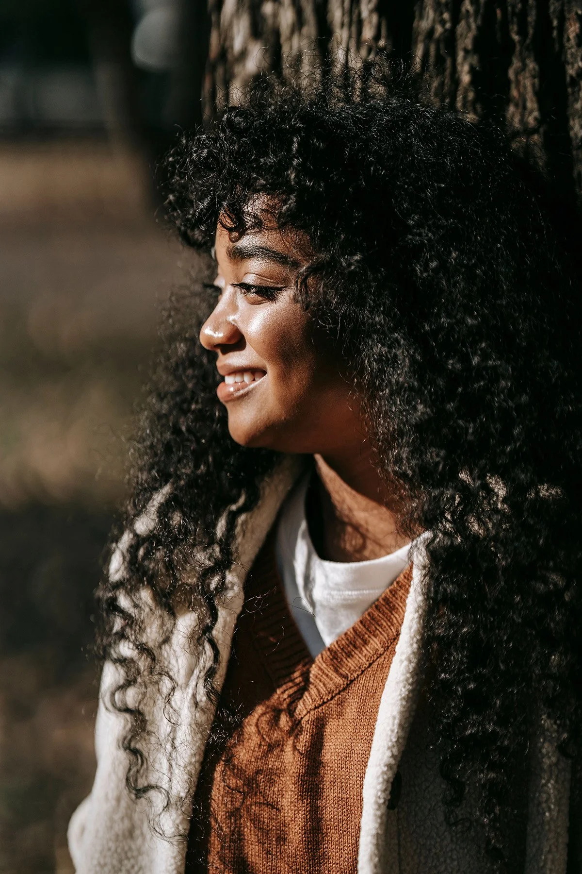 A side profile of a smiling woman with curly black hair, wearing a white t-shirt, brown sweater, and beige cardigan, standing outdoors in front of a tree trunk. Toronto Therapy