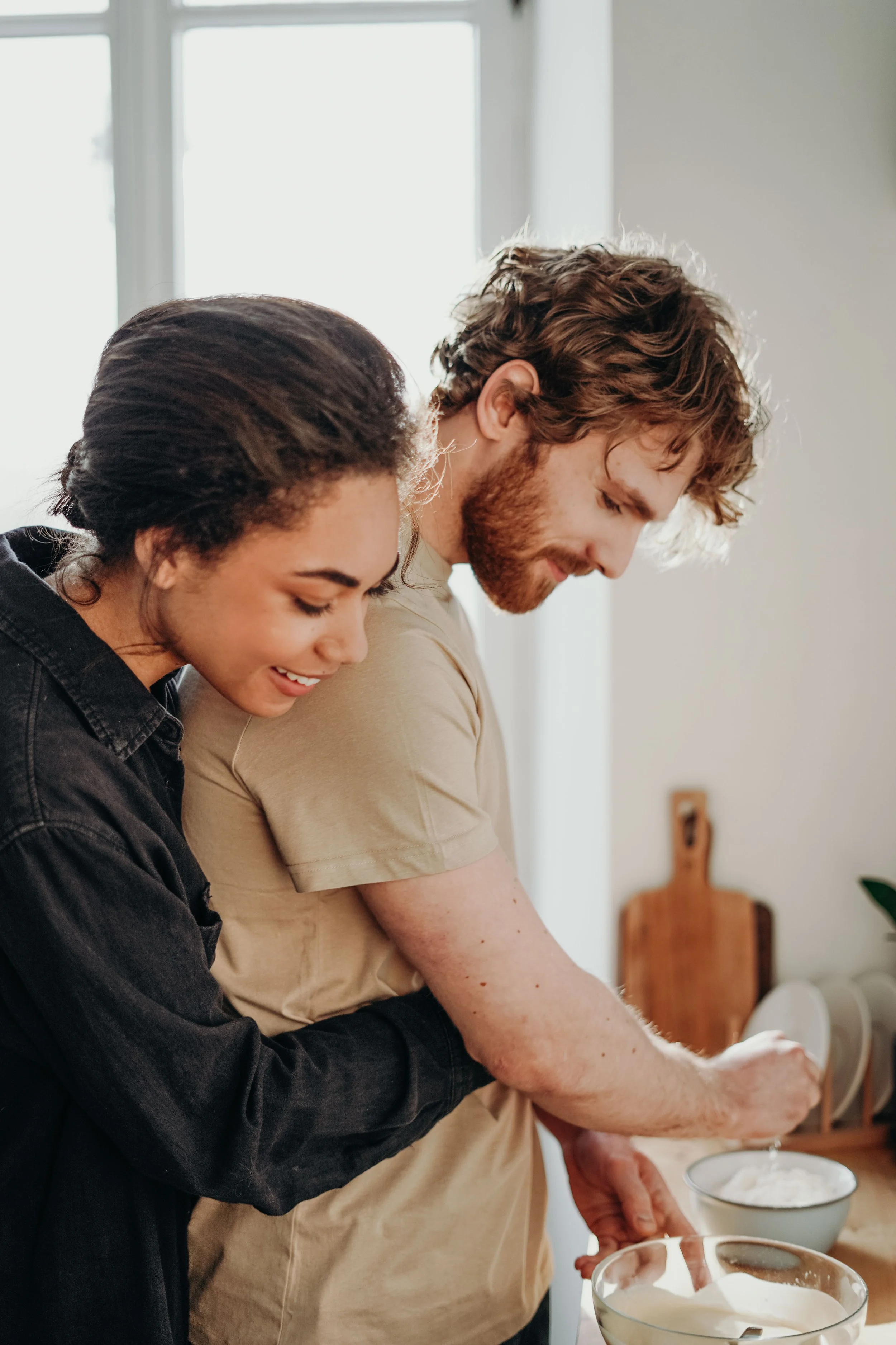 Two people, a woman with dark curly hair and a man with light brown hair and beard, are standing close together in a kitchen, smiling and preparing food. There are bowls of whipped cream on the counter.