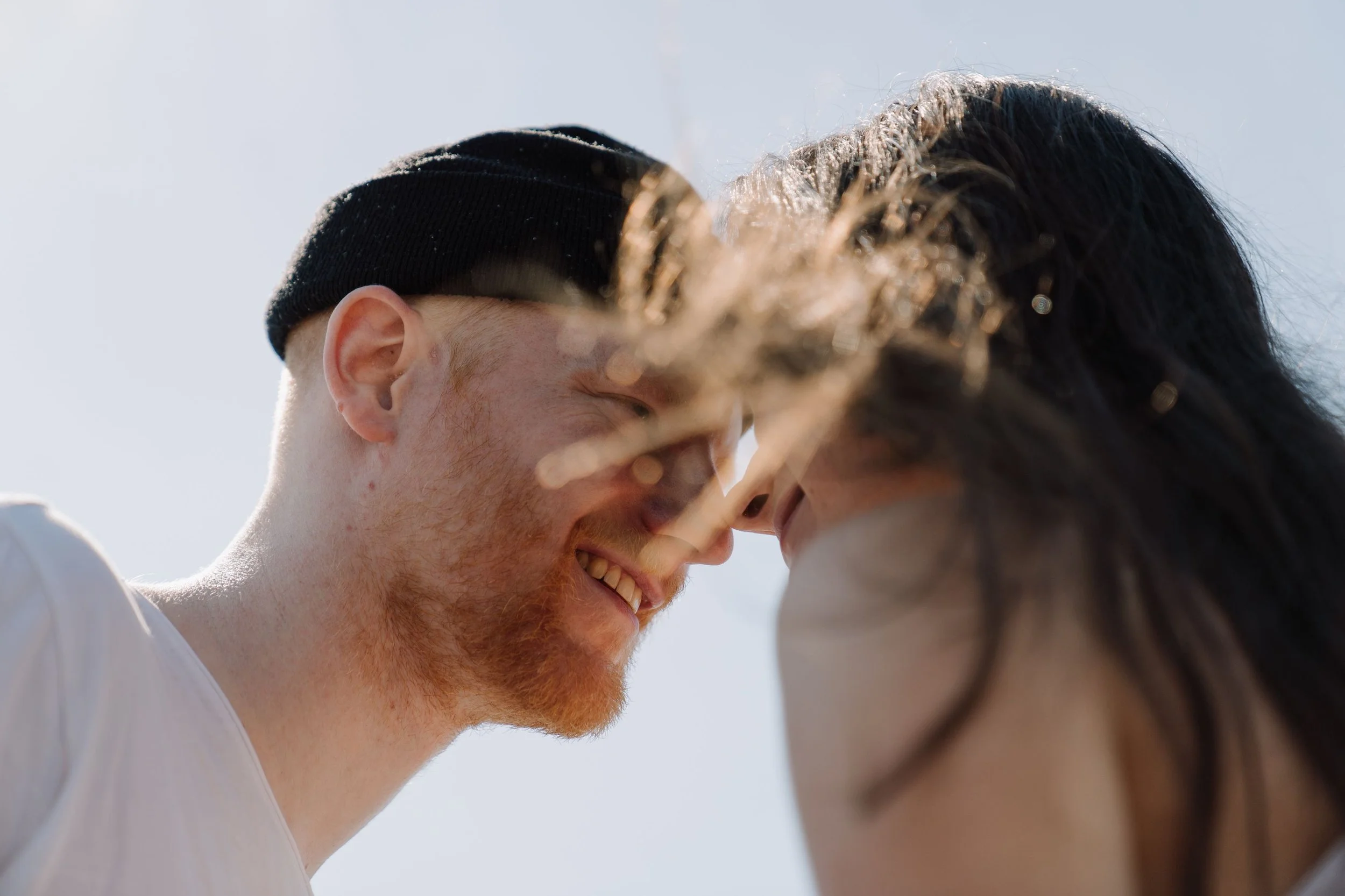 A close-up of two people, one with light skin and red hair wearing a black beanie, and the other with dark hair and skin tone, leaning in close, sharing a tender moment outdoors.