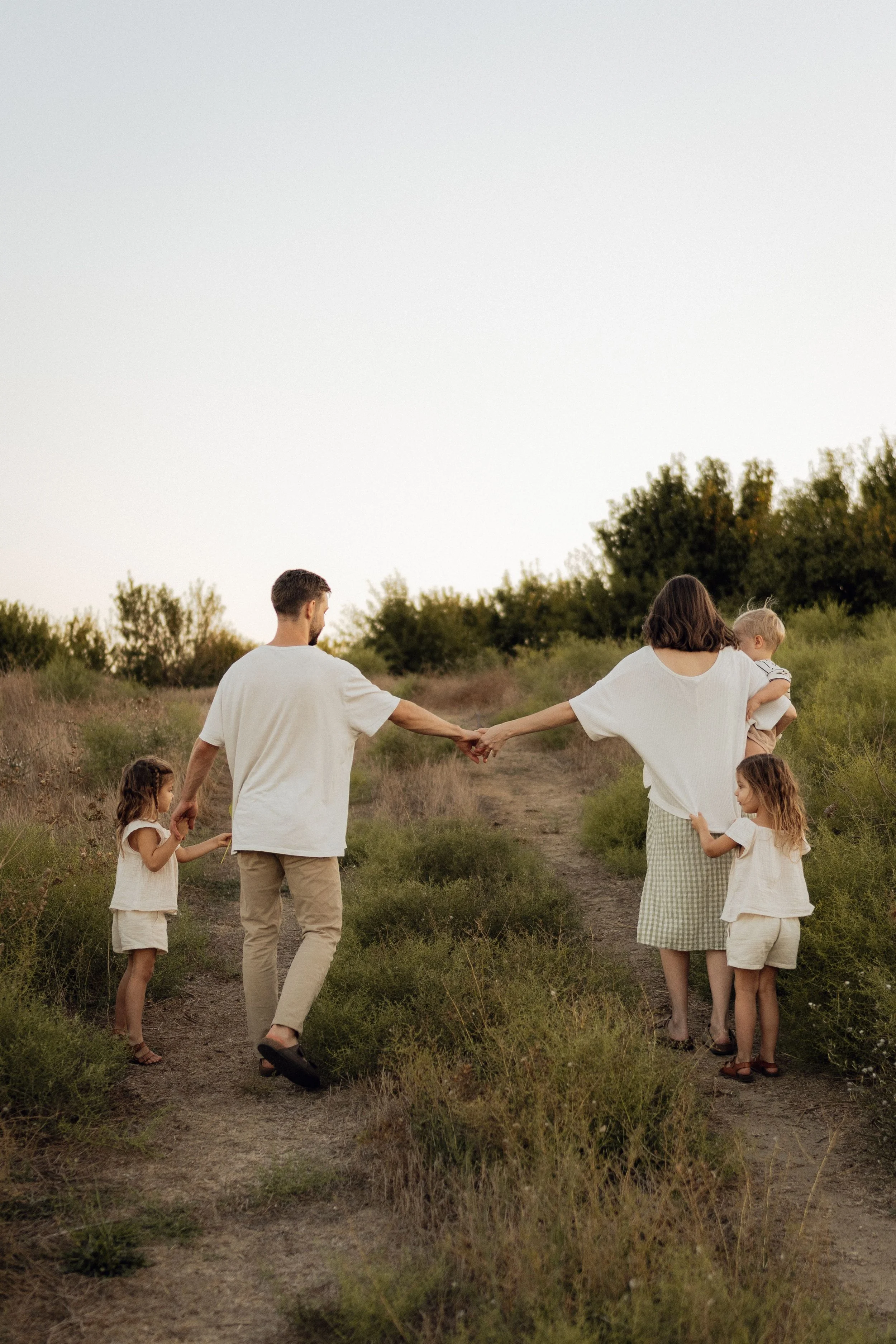 A family of six walking hand-in-hand along a dirt path through a grassy field with trees in the distance during sunset. Noema Therapy in Toronto, Ontario
