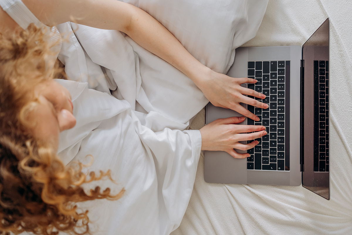 Person with curly red hair using a silver laptop while lying in bed on white bedding.