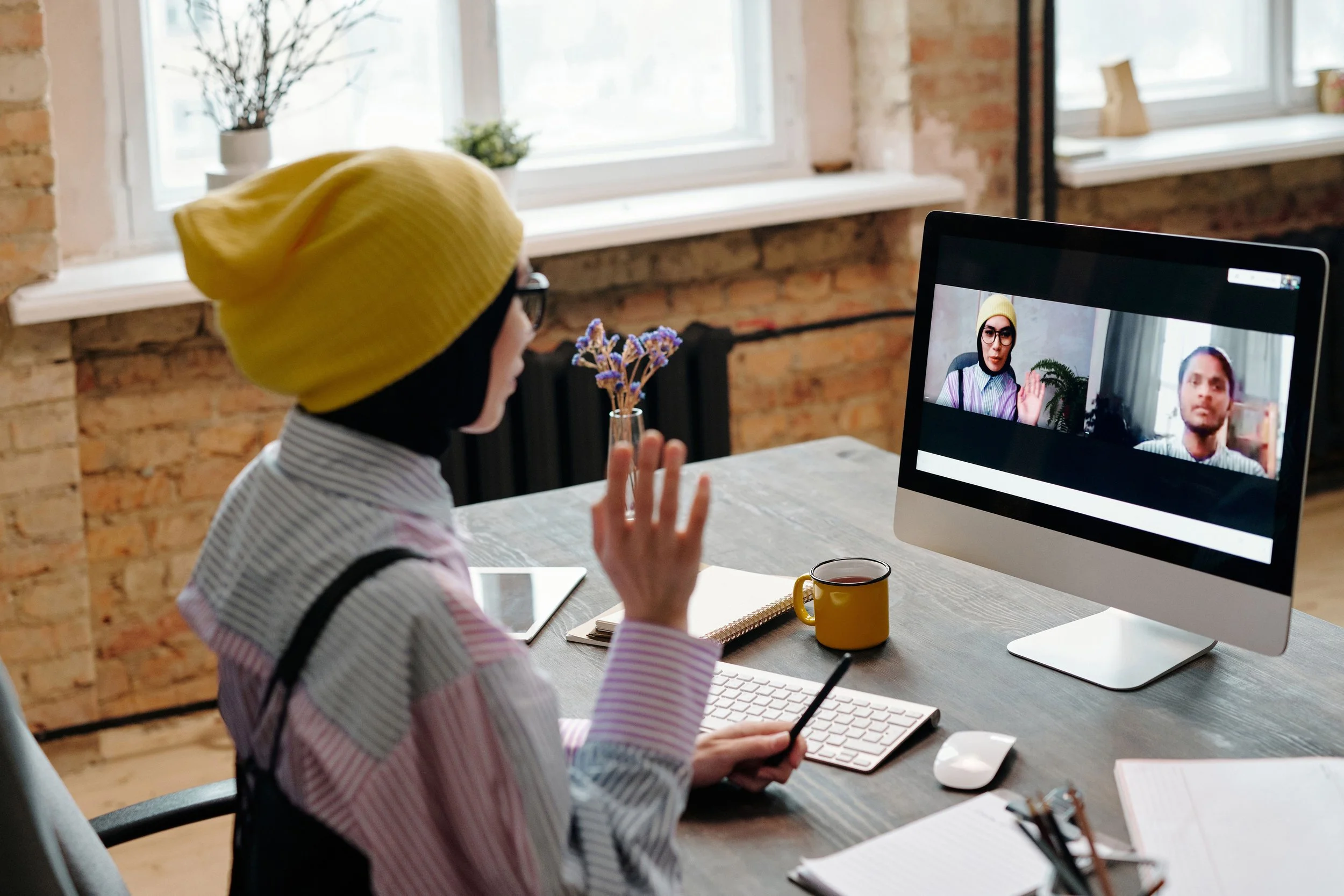 A person wearing a yellow knit hat and glasses participating in a video conference call on a desktop computer in a well-lit room with brick walls, plant decorations, and a coffee mug on the desk. Couples Therapy