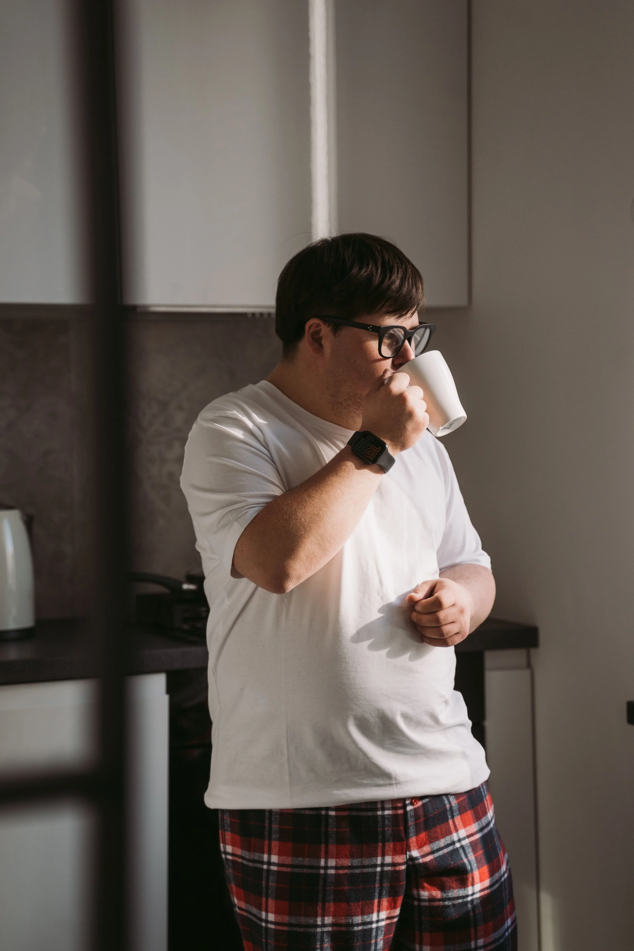 A man with dark hair and glasses drinking from a white mug in a kitchen, wearing a white t-shirt and red plaid shorts. Therapy in Toronto ontario