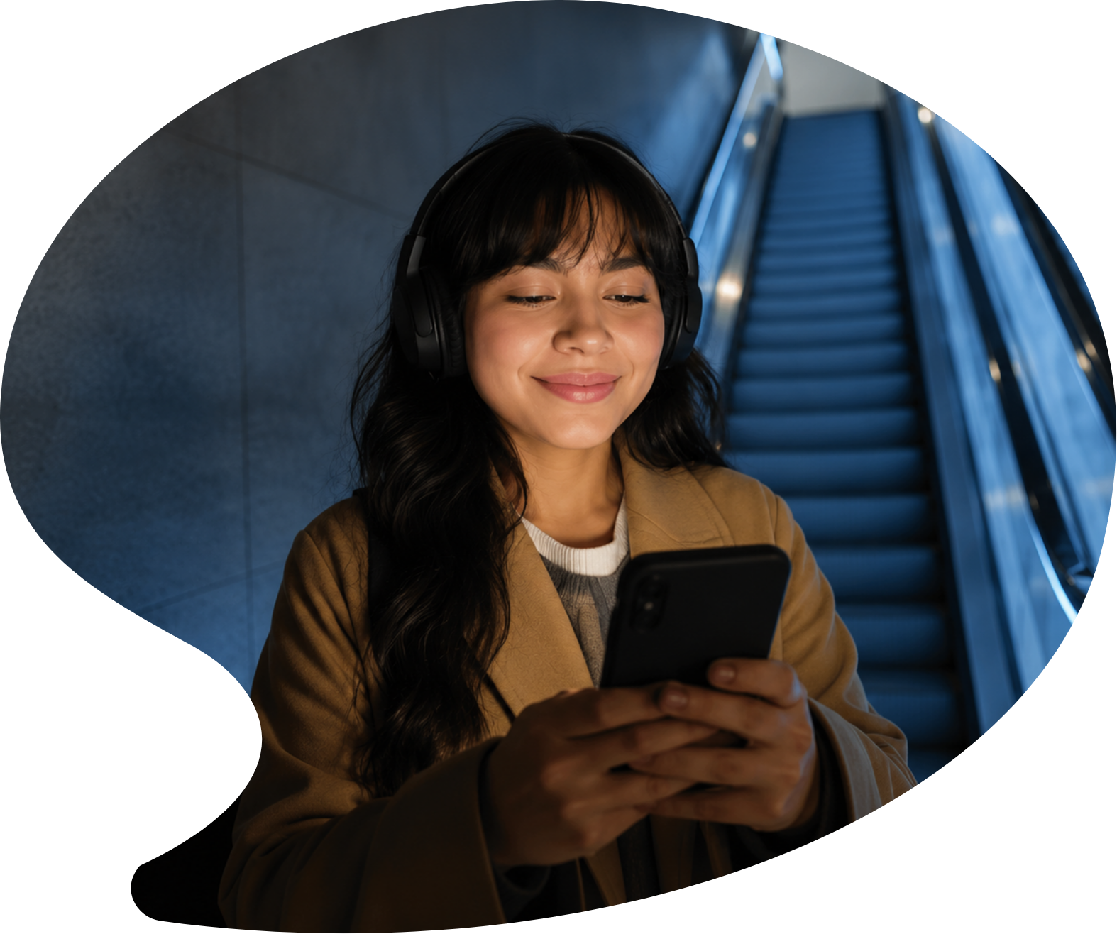 Young woman with dark hair wearing headphones and a tan coat, looking at her smartphone, in front of an escalator in a modern indoor space.