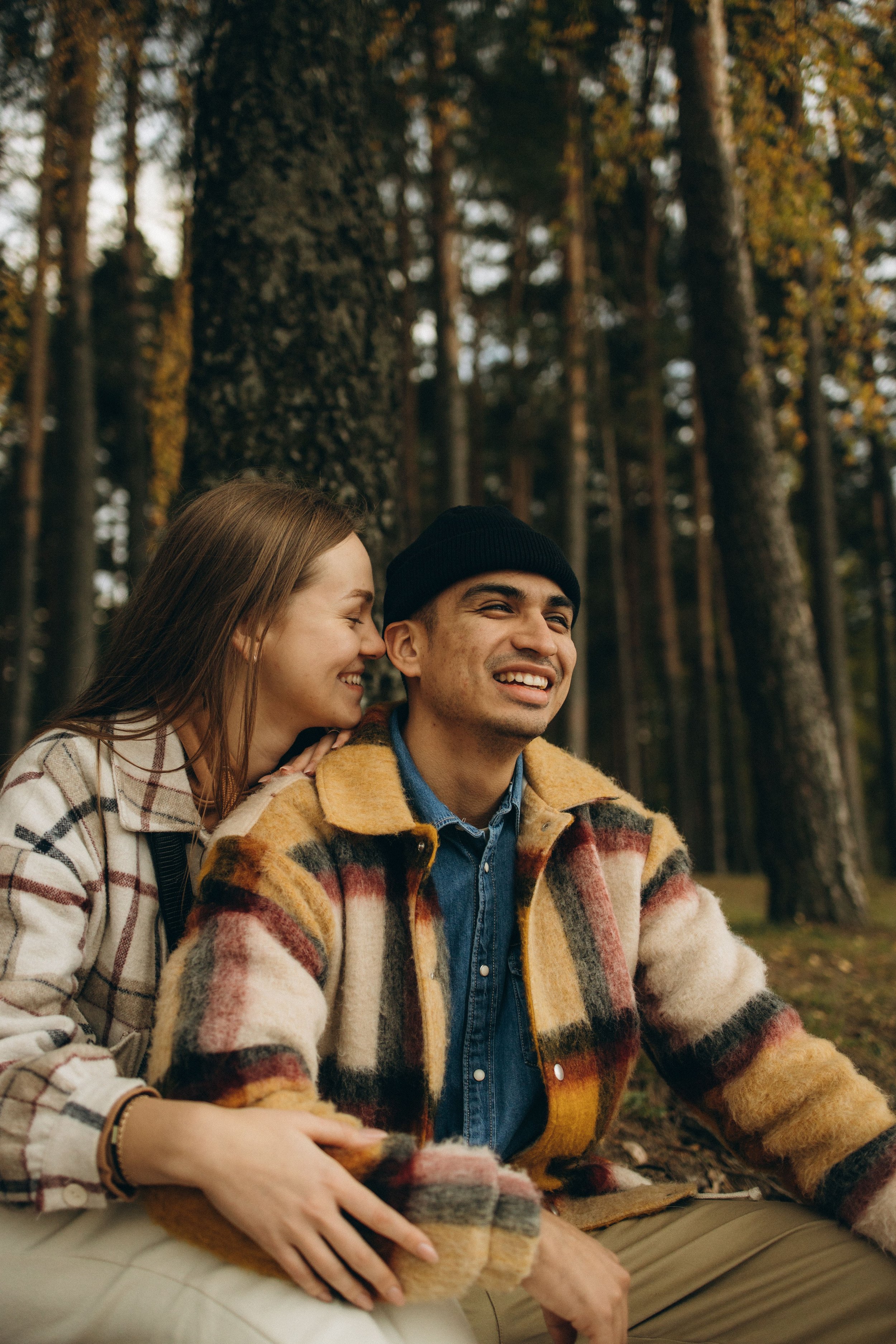 A smiling couple sitting outdoors in a forest, sharing a happy moment, wearing plaid jackets suitable for cool weather.