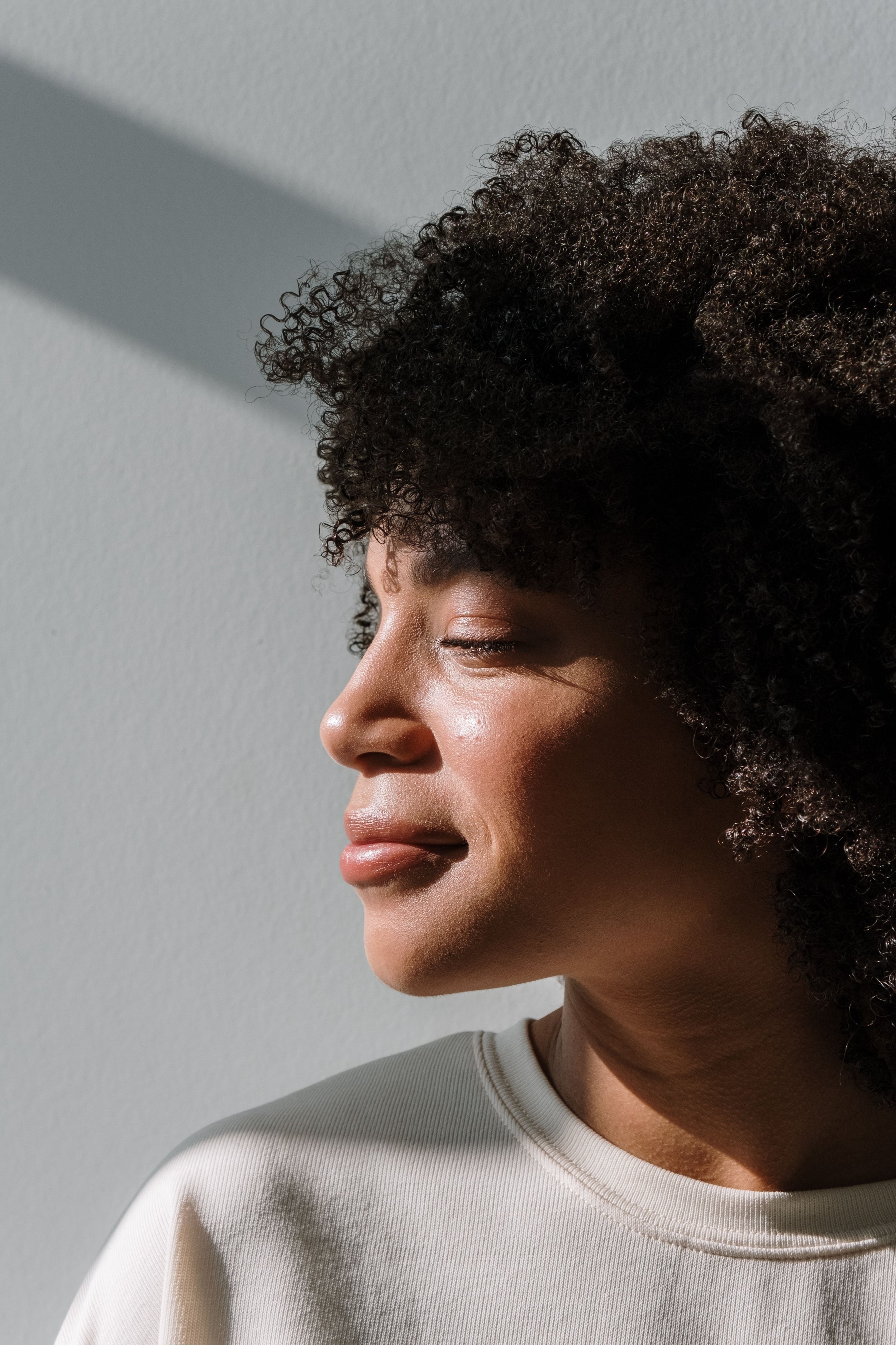 Close-up of a woman with natural curly hair, minimal makeup, and a light-colored top, standing against a plain wall with shadows.
