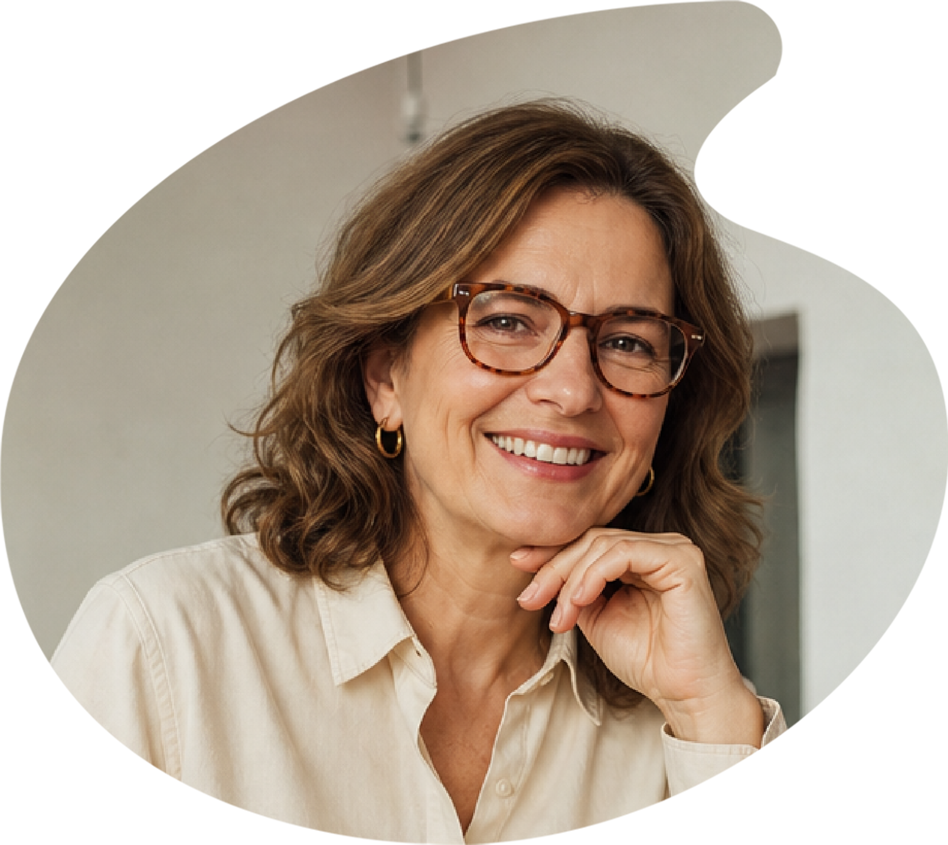 A smiling woman with brown hair and glasses resting her chin on her hand, sitting in a room with a white wall.
