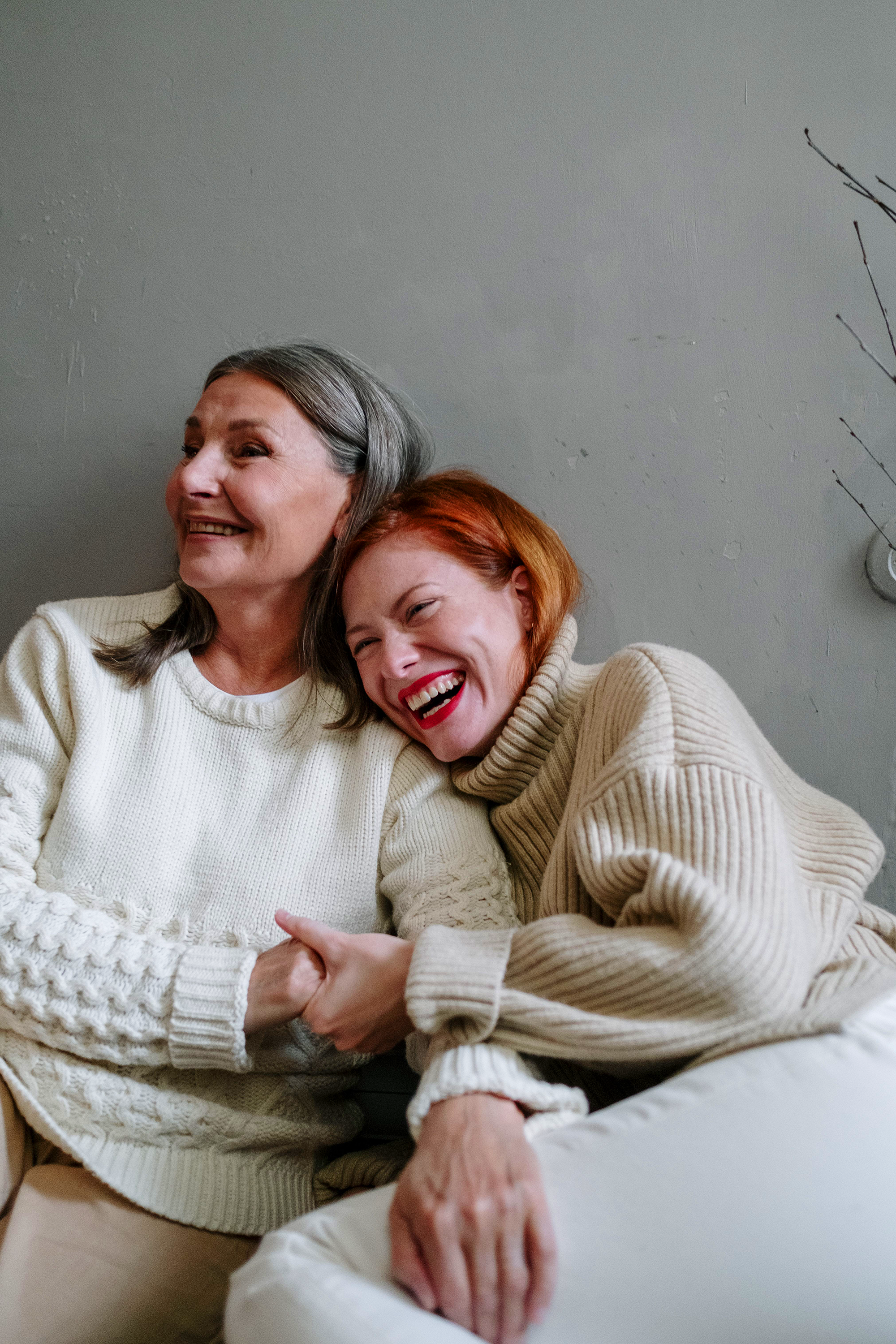 An elderly woman with gray hair and a young woman with red hair sharing a warm hug, both smiling, sitting on a bed in a cozy room.