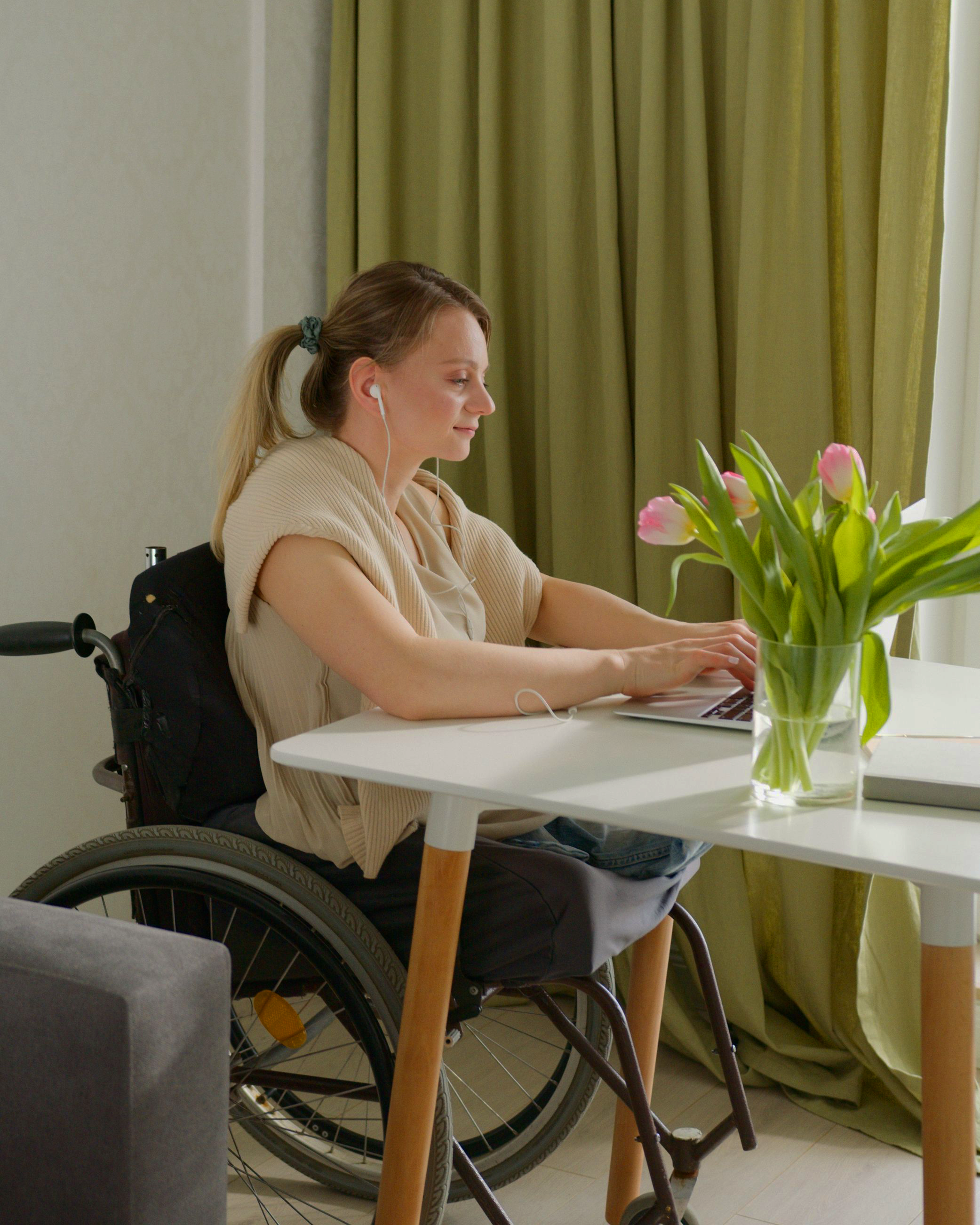 A woman with light skin, brown hair tied back, wearing a beige shirt and white earphones, sitting in a wheelchair at a white table with a laptop in front of her, working indoors. There is a vase with pink tulips on the table, a green curtain in the background, and a window to her right.