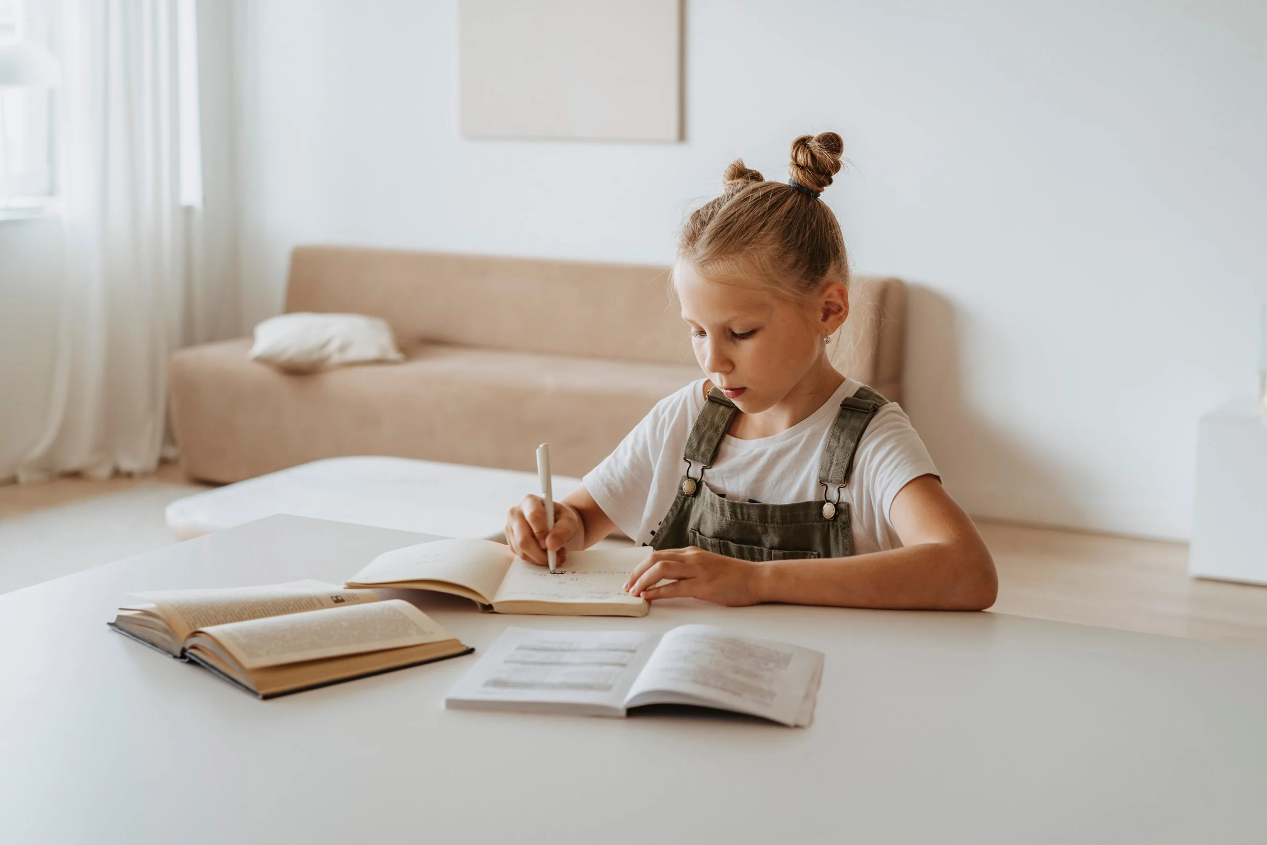 A young girl with hair styled in two buns is sitting at a white table in a bright room, writing in a notebook. Several open books are spread out on the table in front of her, and she appears focused on her writing.