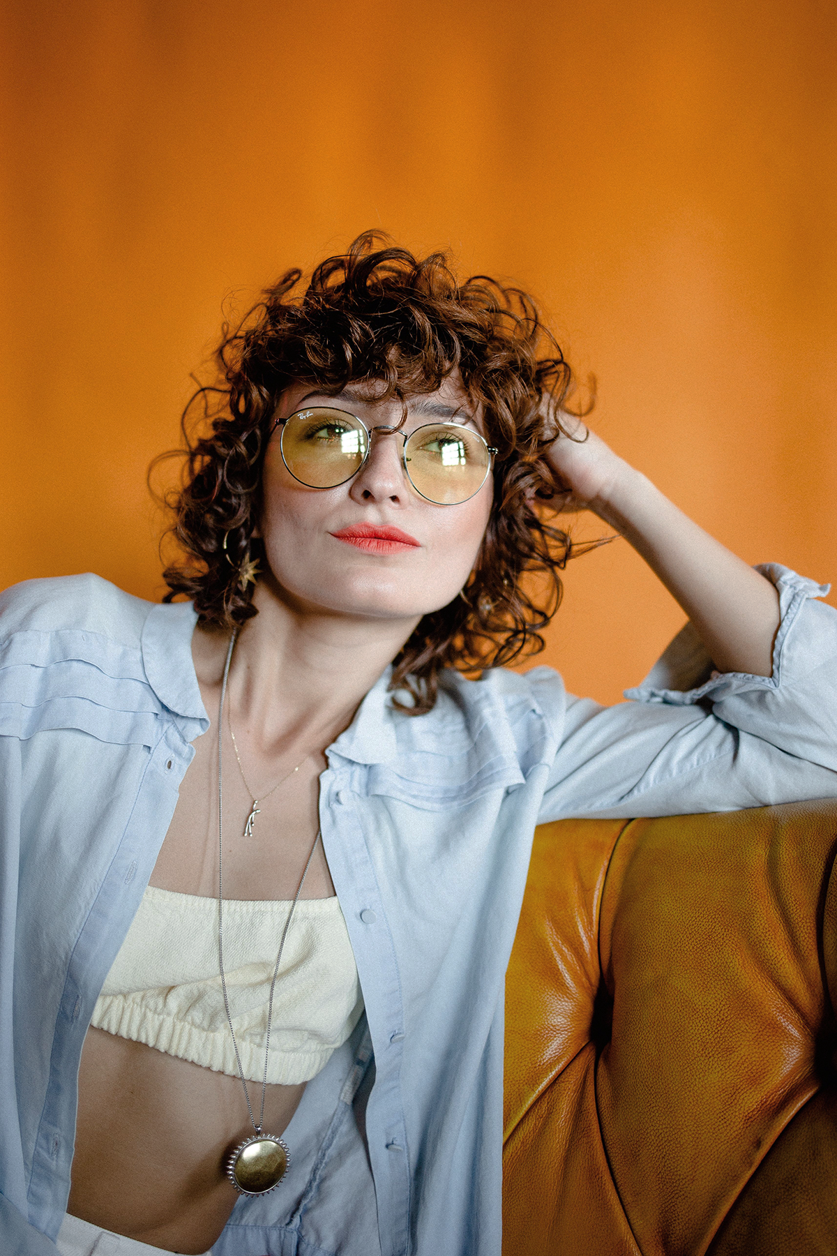 A woman with curly red hair wearing yellow glasses, a white top, and a light blue button-up shirt, sitting on a brown leather sofa against an orange background.